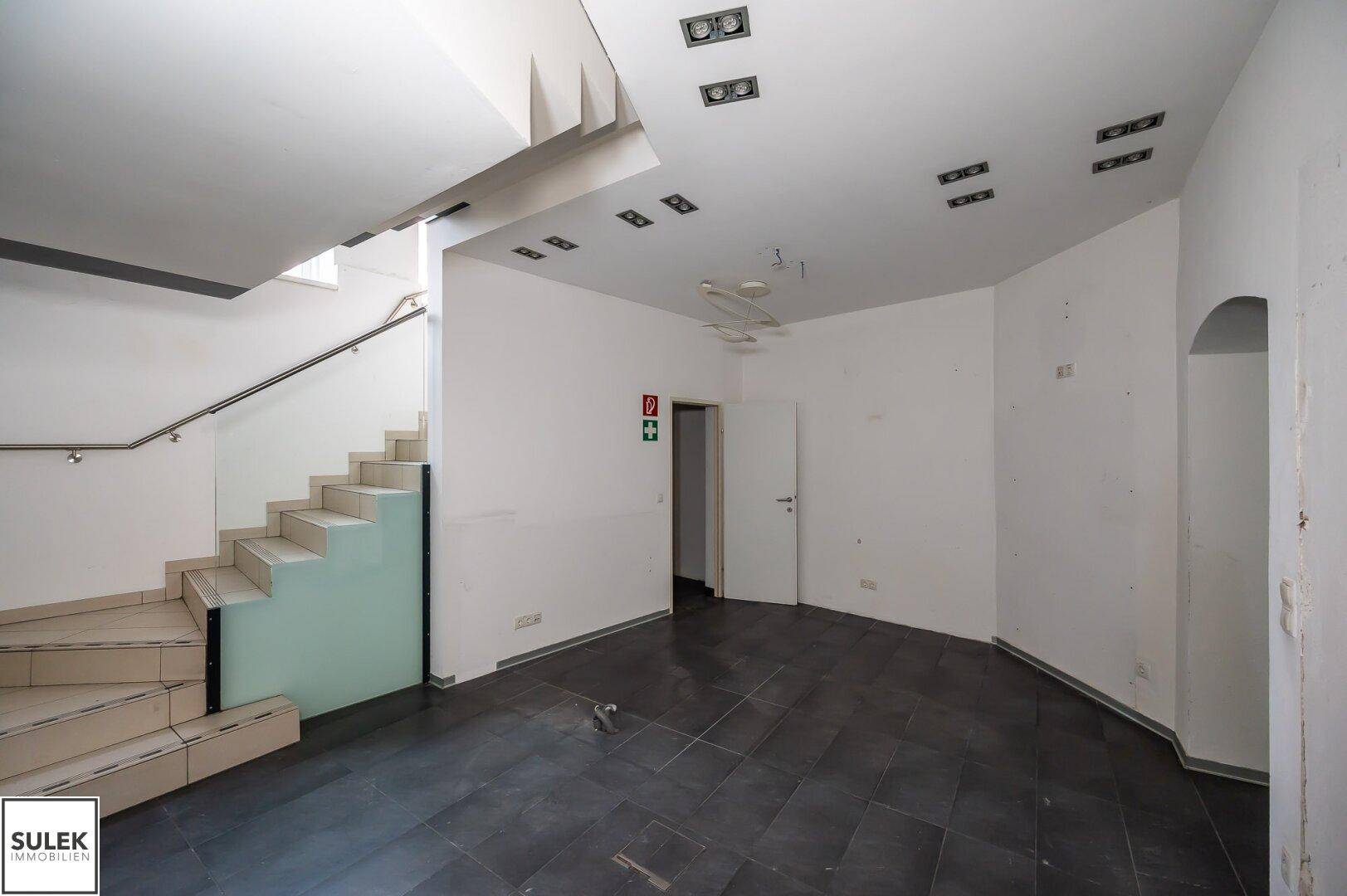 Geräumiger Büroraum mit dunklen Fliesen, weißen Wänden und einer Treppe mit Glasgeländer.