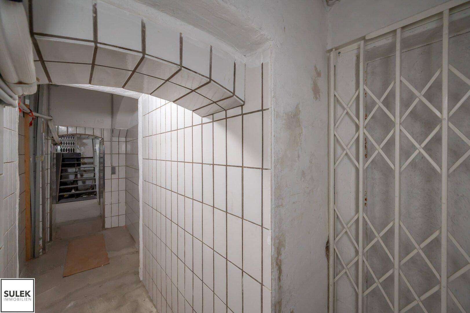 Gefliester Kellergang mit Gittertür und gewölbter Decke, der zu weiteren Kellerräumen führt.
