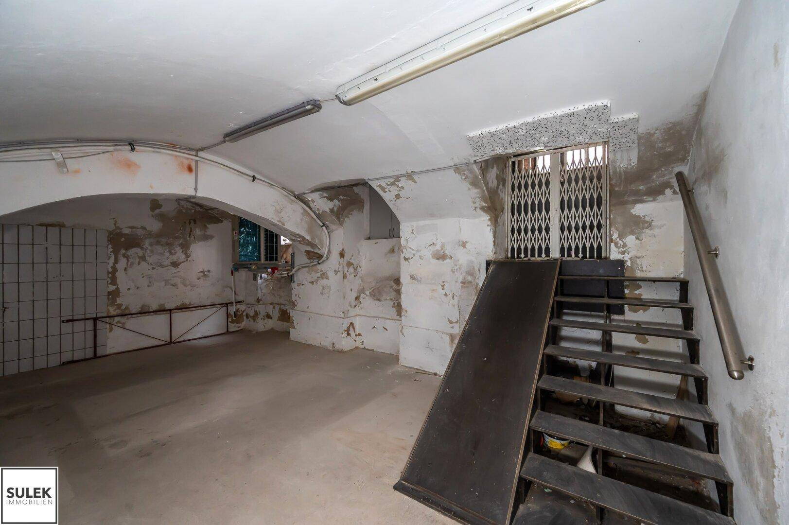 Großer Kellerraum mit Rampe und Treppe, gefliesten Wänden und Gittertür, der Renovierungsbedarf zeigt.