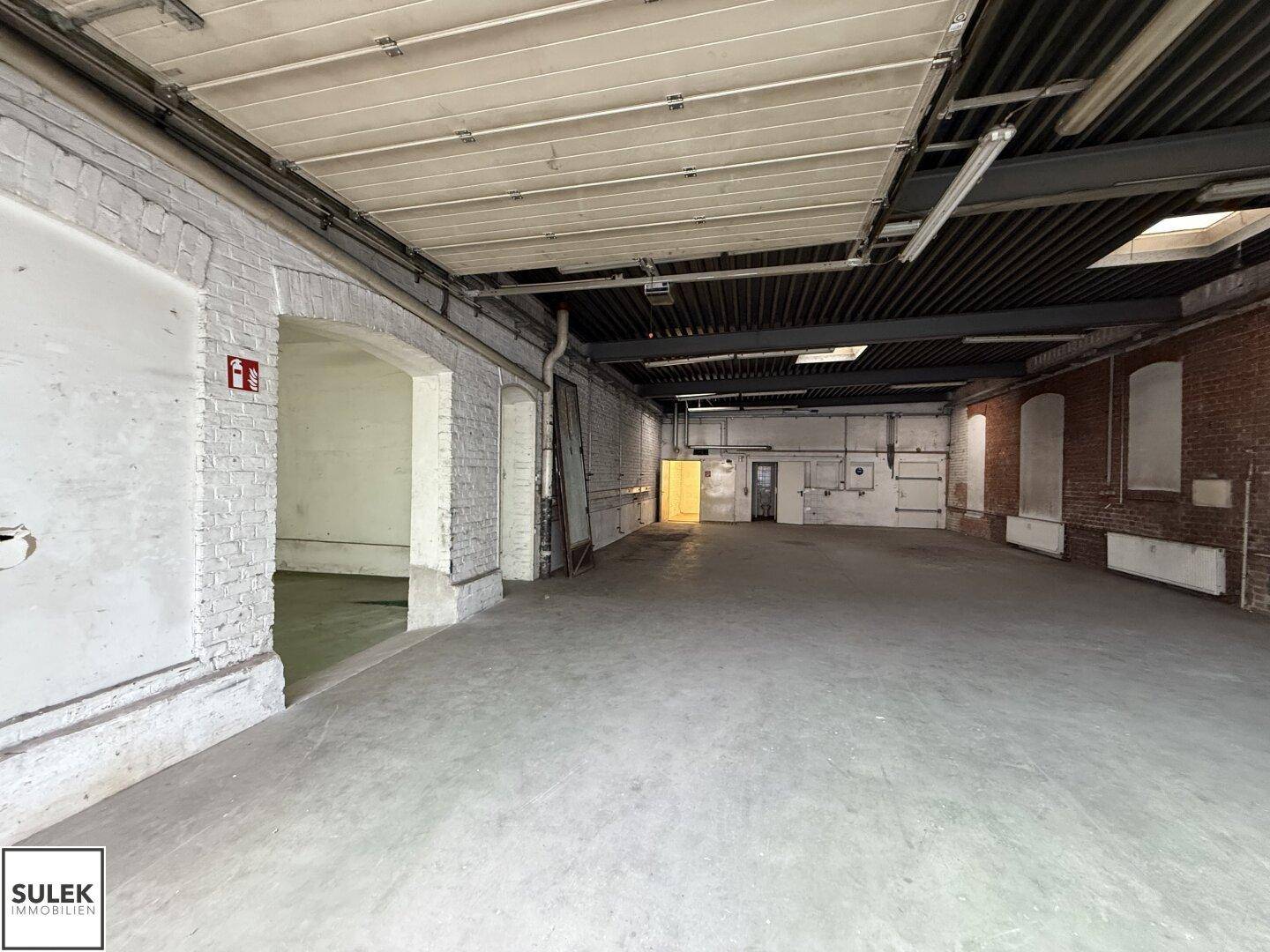 Großer, leerer Lagerraum mit hohen Decken, Betonboden und sichtbaren Ziegelwänden.