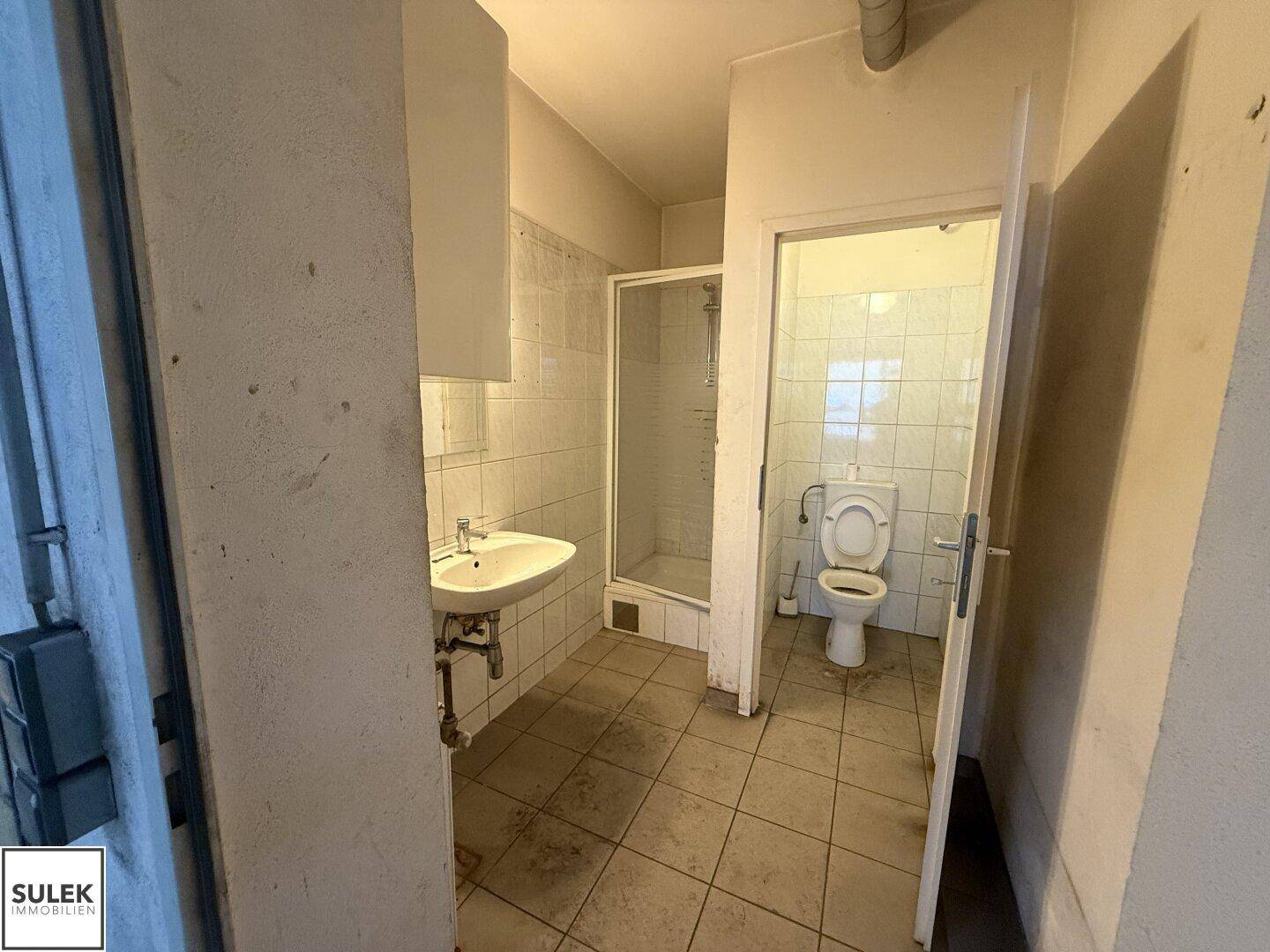 Einfaches Badezimmer mit Duschkabine, Waschbecken und Toilette, gefliest in Weiß und Beige.