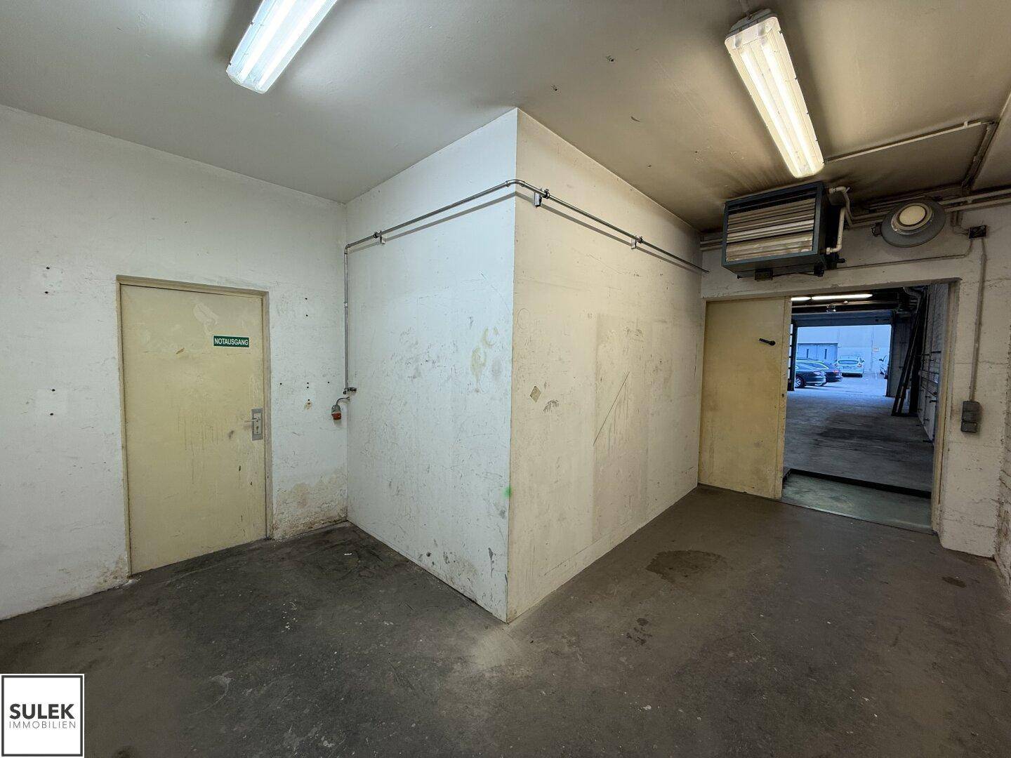Kleinerer Raum mit zwei Türen und einem Heizlüfter an der Wand, mit Betonboden.