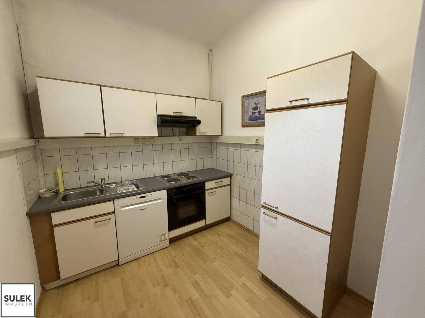 Kleine Einbauküche mit weißen Schränken, Spüle, Herd und Kühlschrank in funktionalem Zustand.