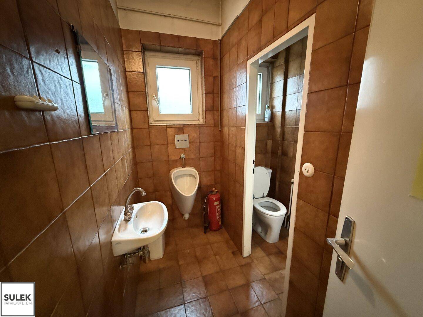 Badezimmer mit braunen Fliesen, Waschbecken, Urinal und Toilette in funktionalem Zustand.