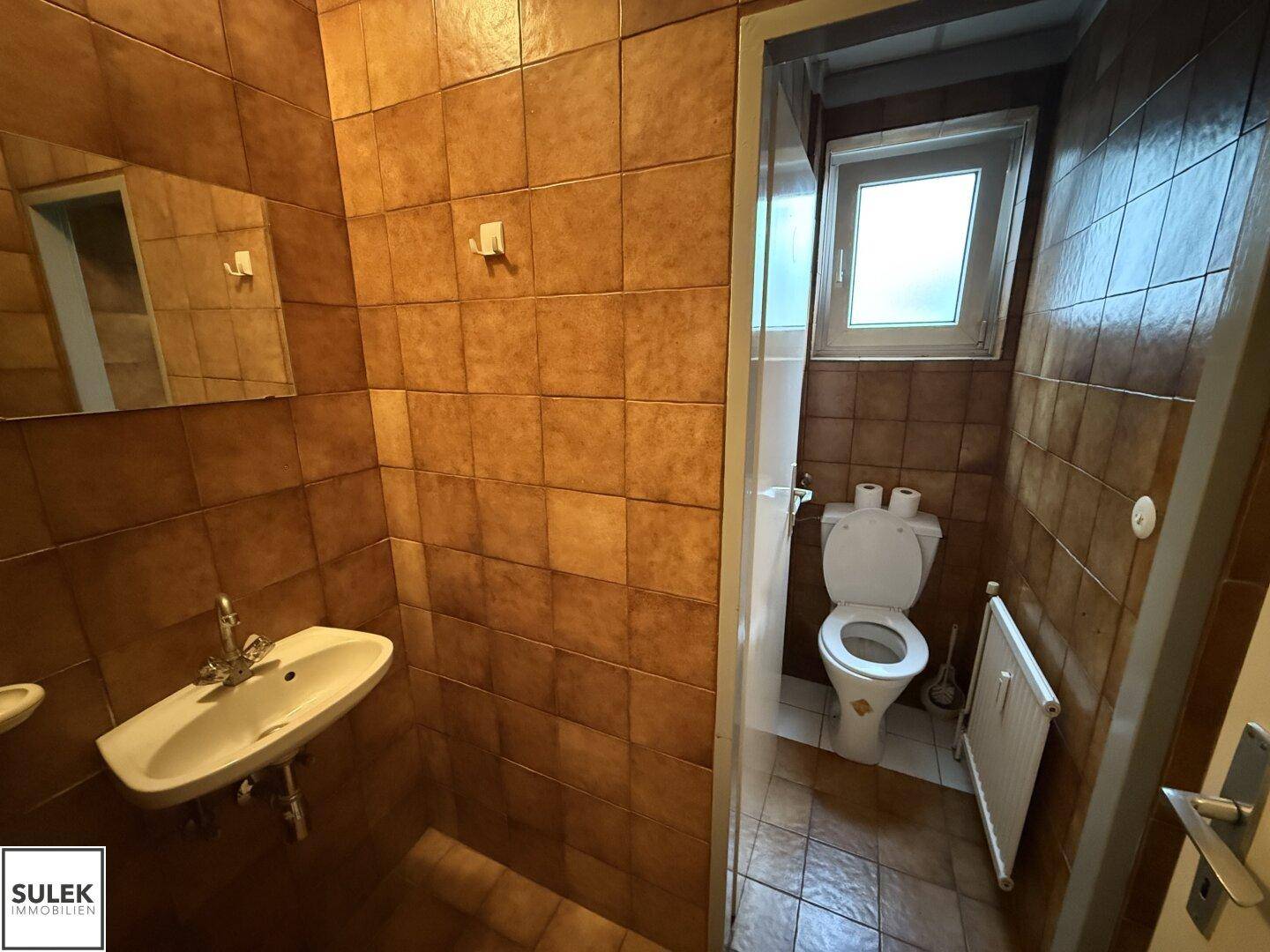 Toilette mit braunen Fliesen, Waschbecken und Fenster, in einem einfachen Zustand.