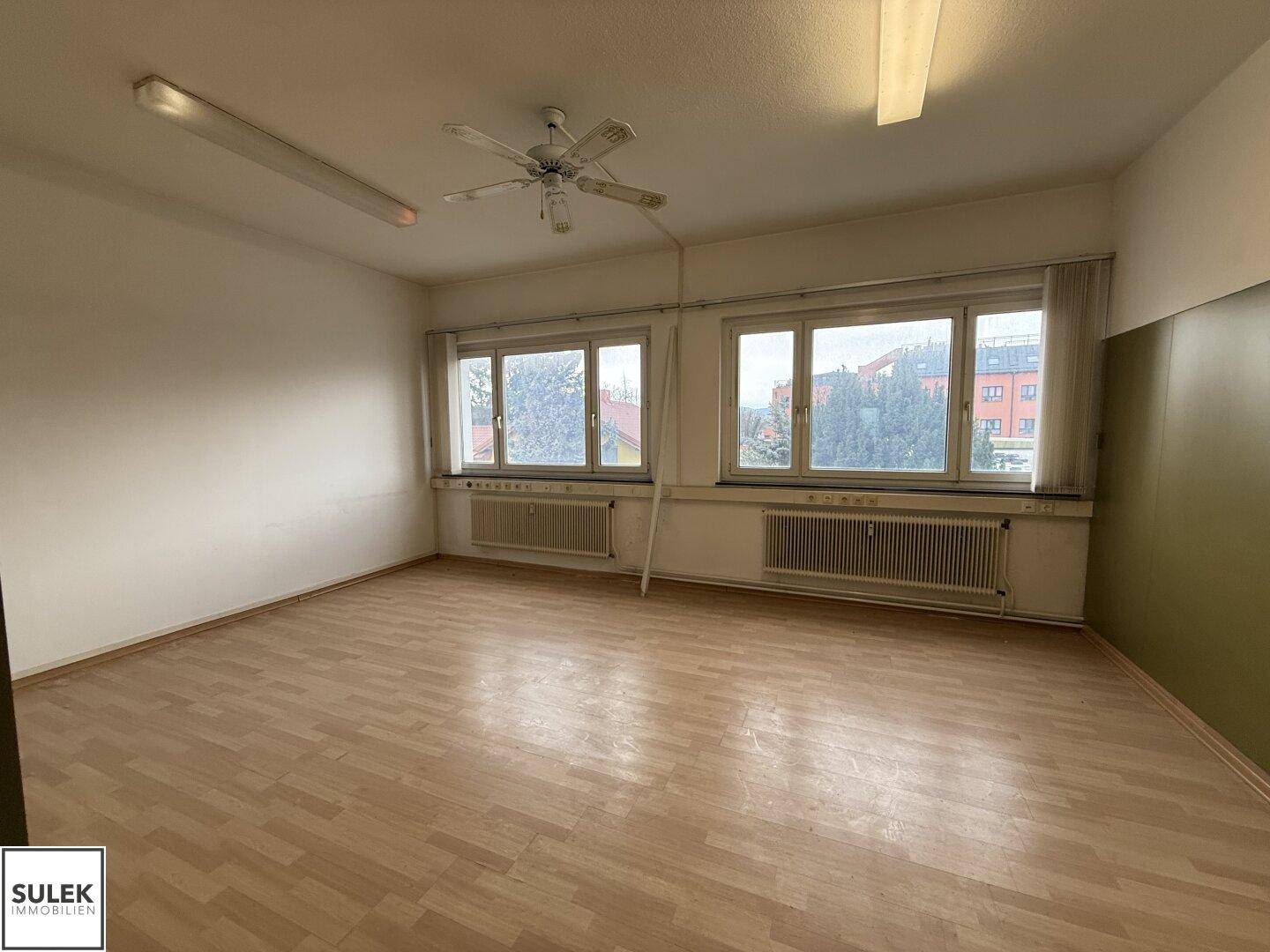Großer Büroraum mit Laminatboden, Deckenventilator und breiten Fenstern mit Blick nach draußen.