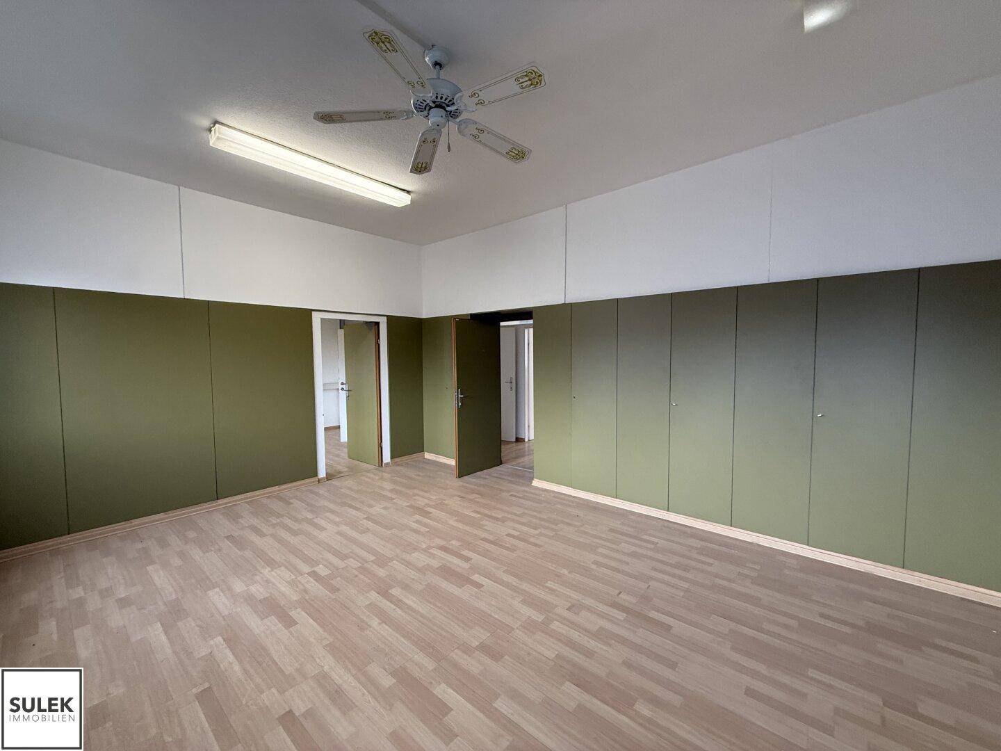 Geräumiger Büroraum mit Laminatboden, Deckenventilator und grünen Einbauschränken entlang der Wand.