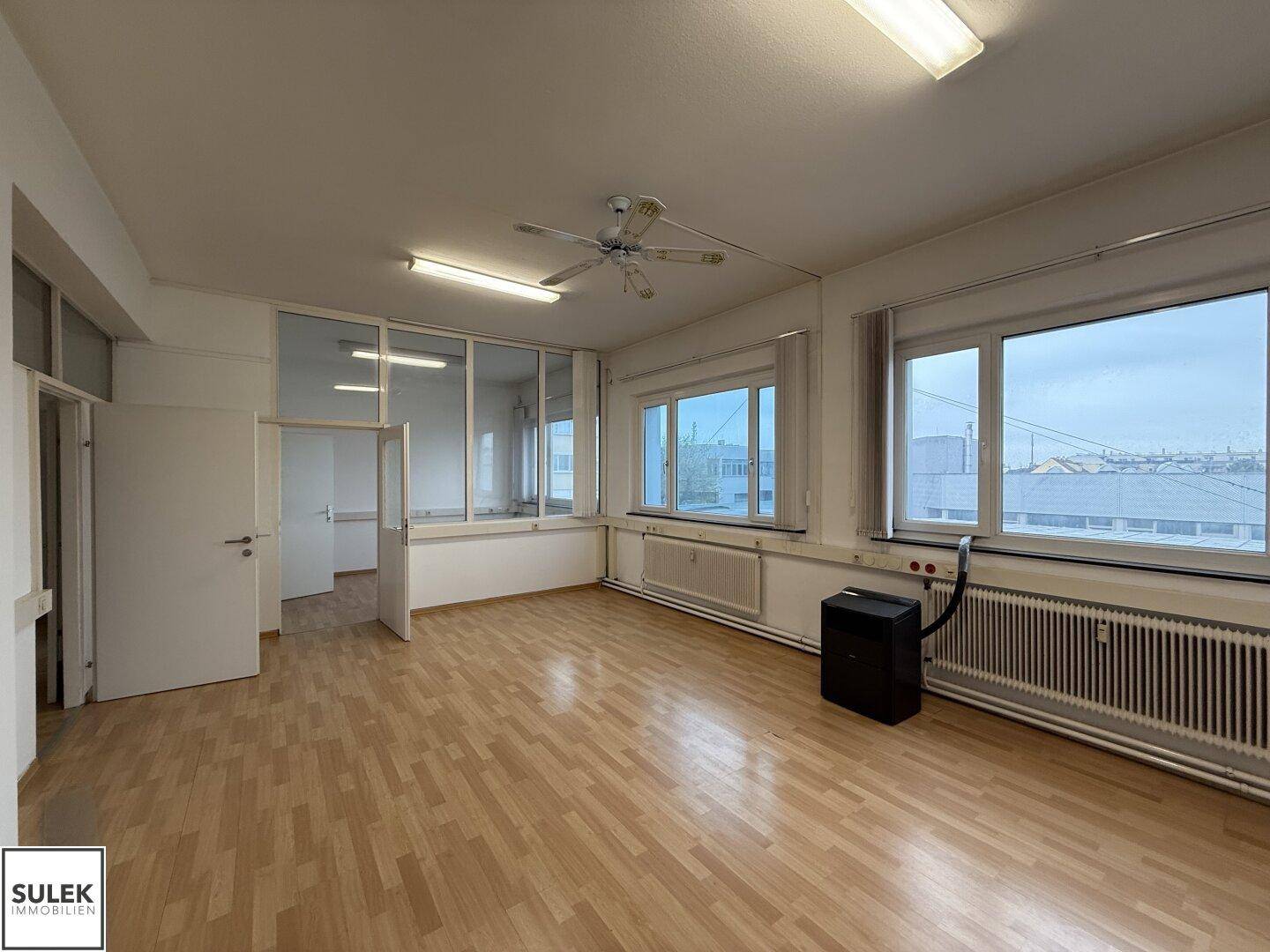 Großer Büroraum mit Laminatboden, Deckenventilator und Glaswänden, die den Raum unterteilen.