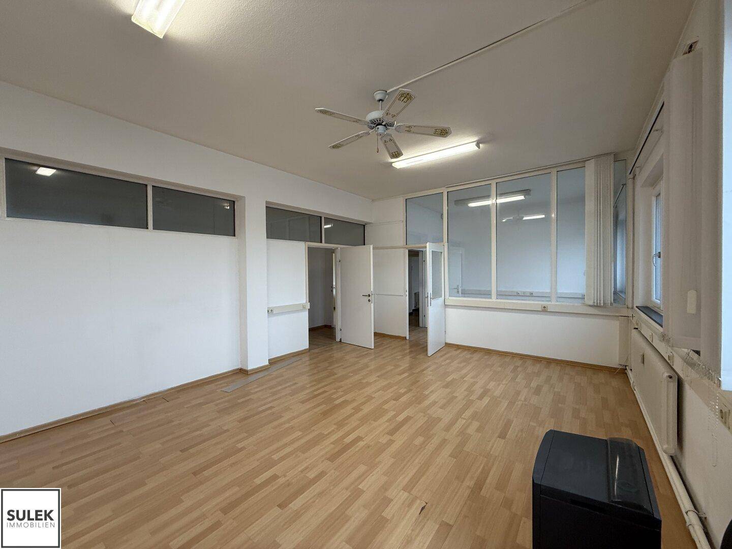 Geräumiger Büroraum mit Laminatboden, Deckenventilator und Glasabtrennungen zu angrenzenden Bereichen.