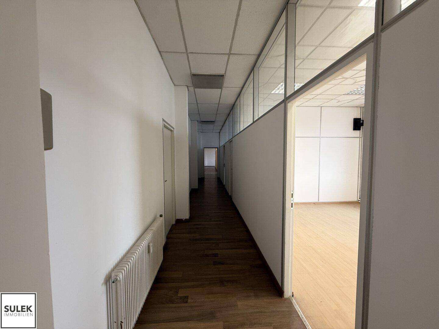 Langer Flur mit Holzboden, weißen Wänden und Glasabtrennungen zu den Büroräumen.