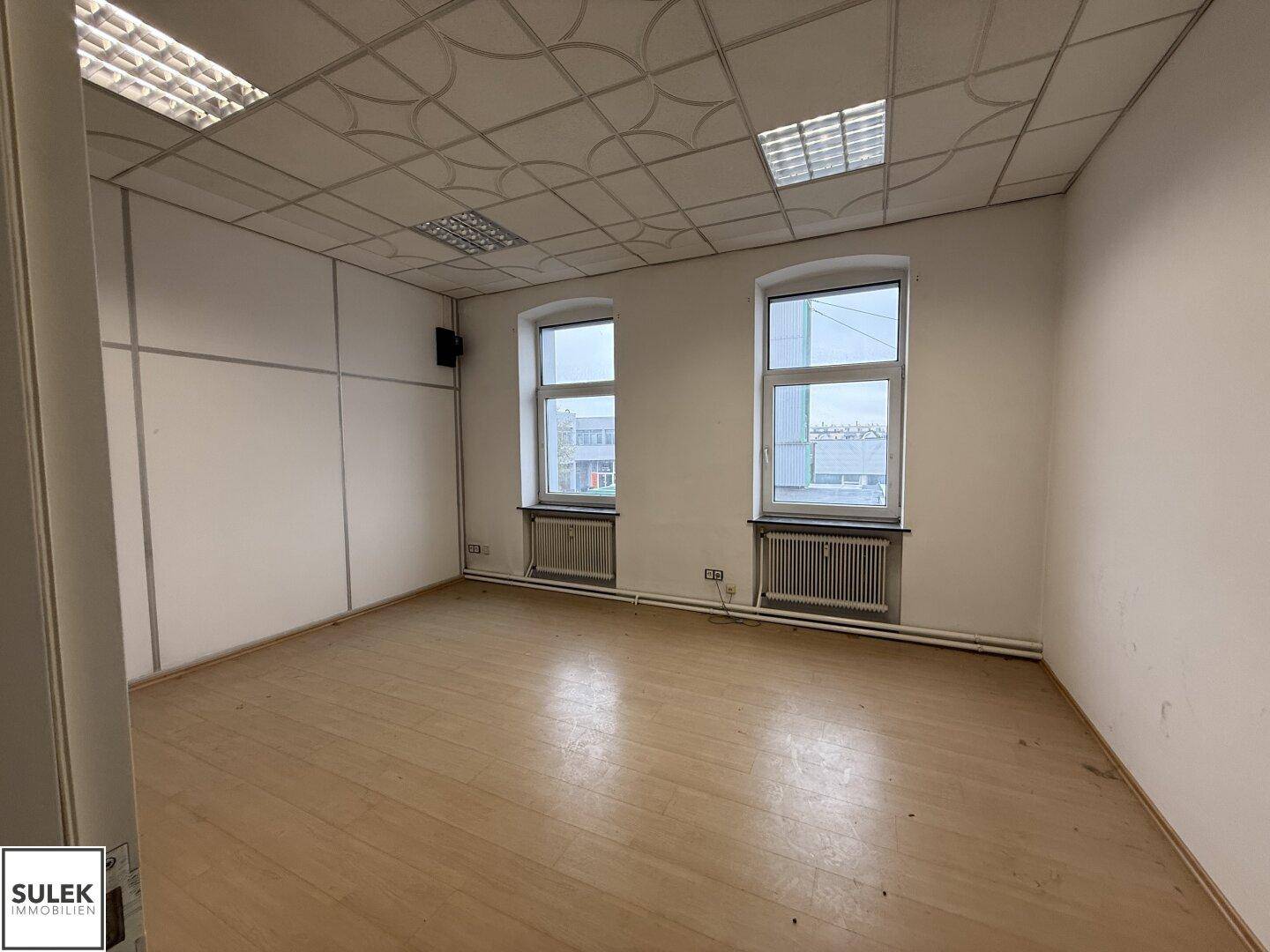 Büroraum mit Laminatboden, zwei Fenstern und einem abgehängten Deckensystem.