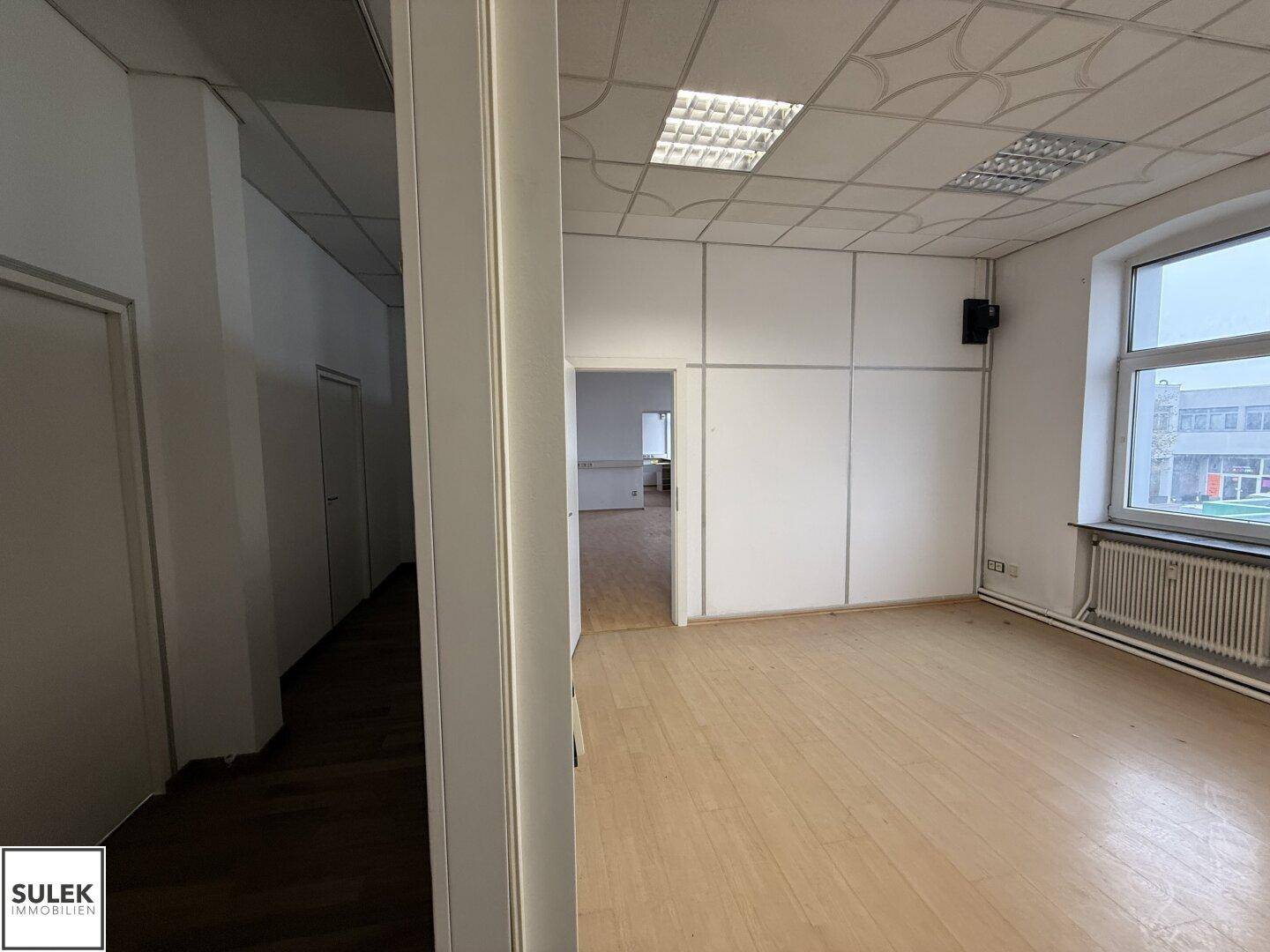 Büroraum mit Laminatboden, Fenster und einer Tür, die zu einem weiteren Raum führt.