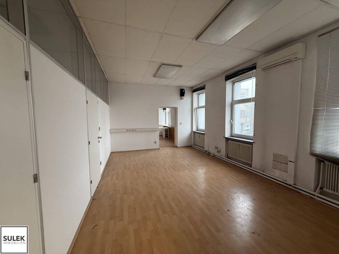 Geräumiger Büroraum mit Laminatboden, Fenstern und Klimaanlage, ideal für mehrere Arbeitsplätze.