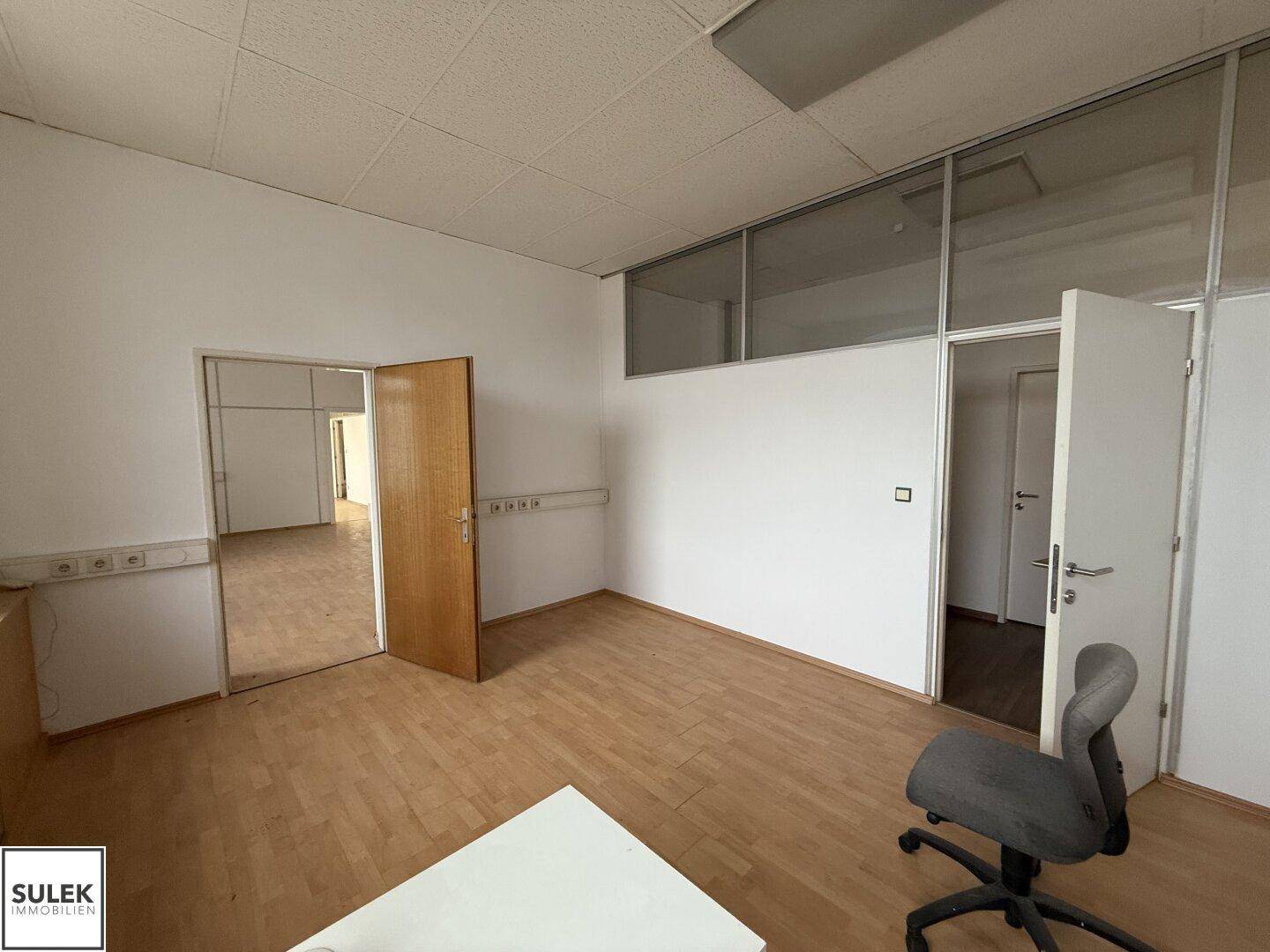 Büroraum mit Holzboden, Fenster und Büromöbeln, mit Blick auf den Flur.