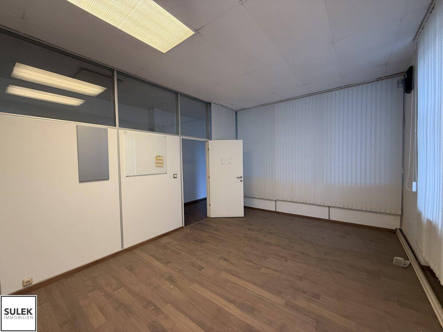 Büroraum mit Holzboden, Fenstern und Jalousien, mit Blick auf den Flur.