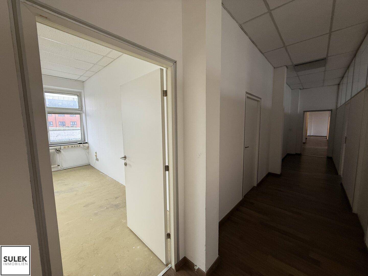 Büroraum mit Holzboden, Fenster und einer Tür, die zu einem weiteren Raum führt.