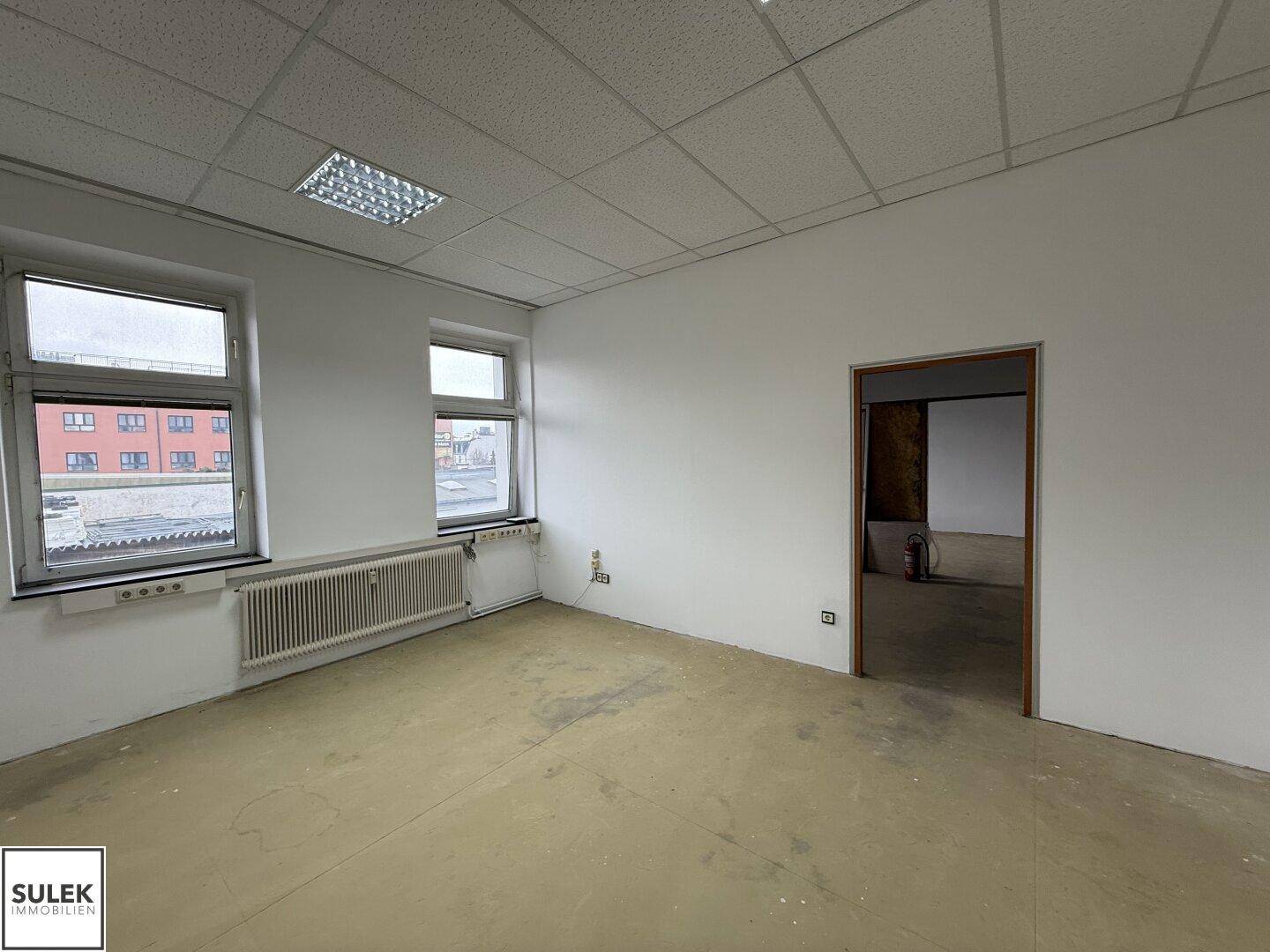 Büroraum mit Holzboden, Fenster und einer Tür, die zu einem weiteren Raum führt.