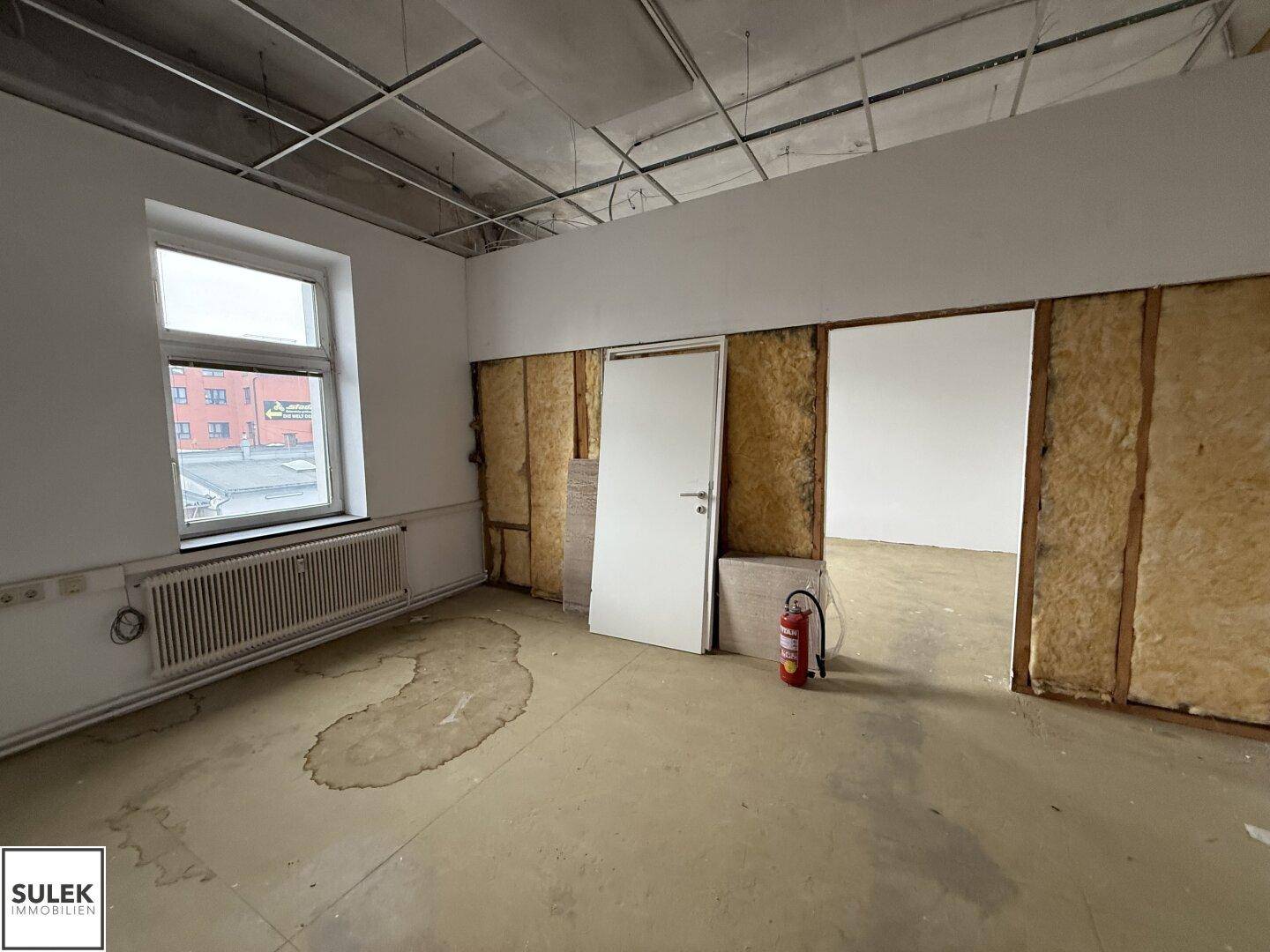 Büroraum mit freiliegender Deckenkonstruktion, Fenster und einem Feuerlöscher.