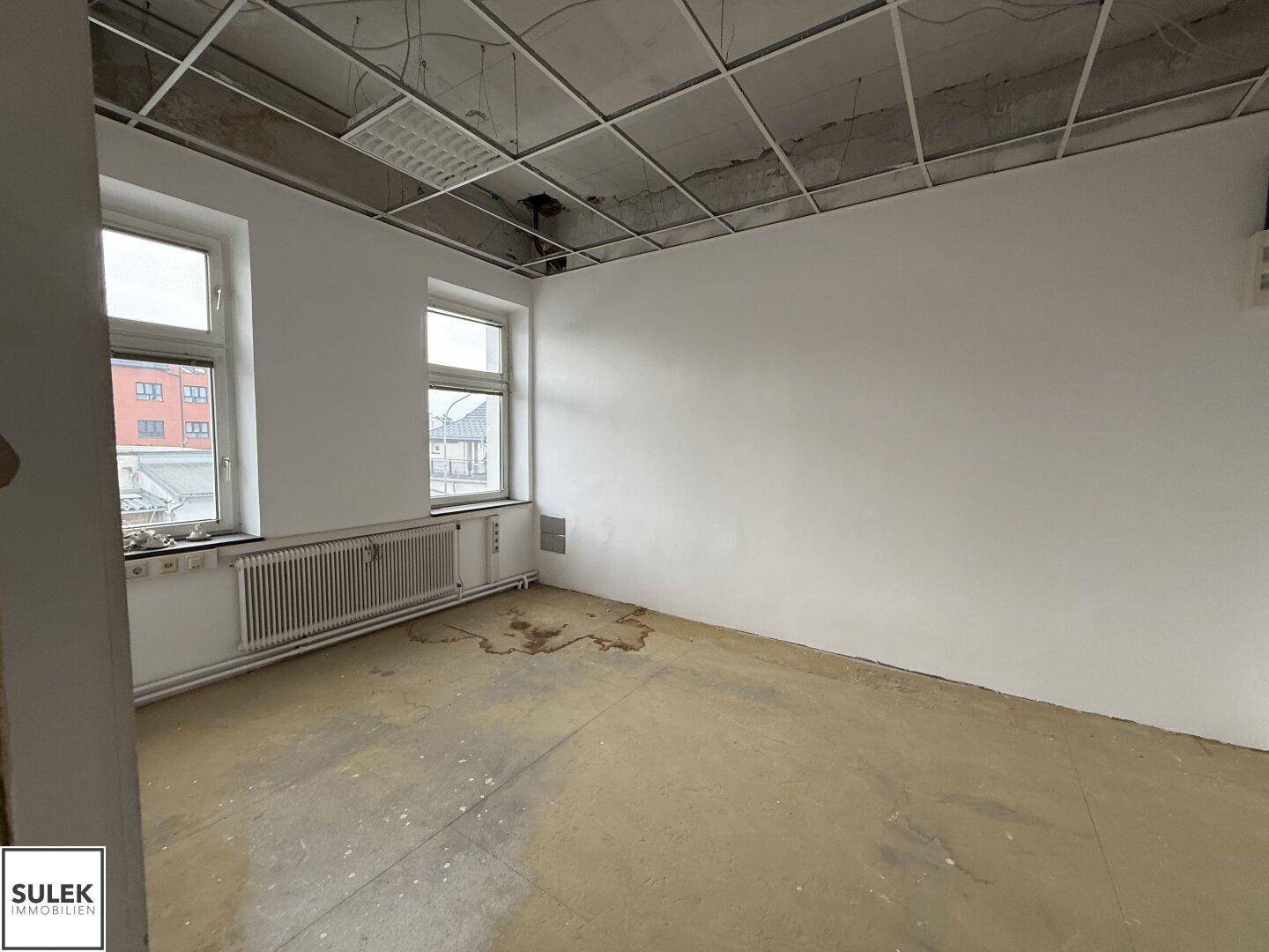 Büroraum mit freiliegender Deckenkonstruktion, zwei Fenstern und einem Heizkörper.