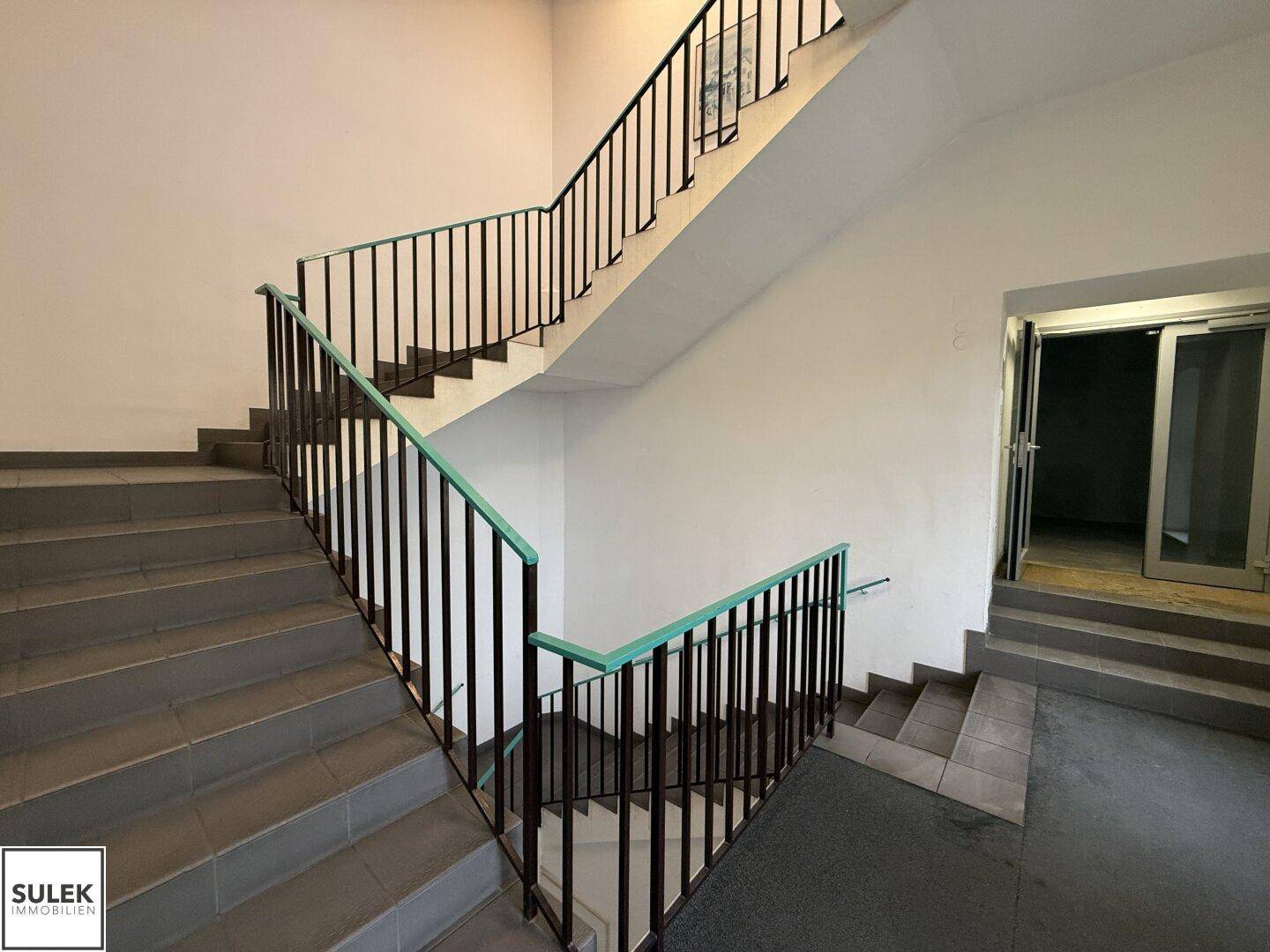 Geräumiges Treppenhaus mit grauen Fliesenstufen und schwarzem Metallgeländer.