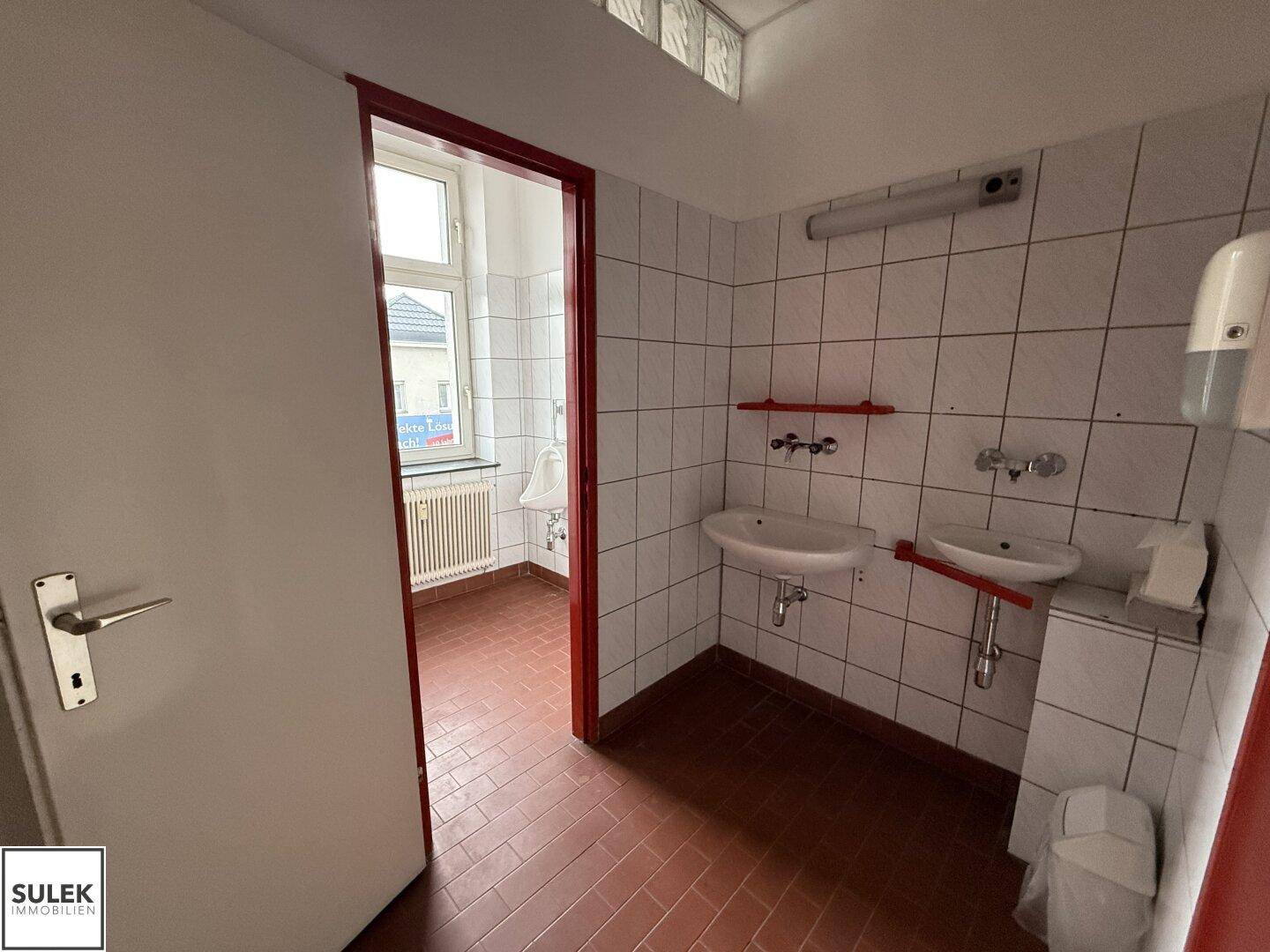 Badezimmer mit roten Fliesen, weißen Wandfliesen, zwei Waschbecken und einem Fenster.