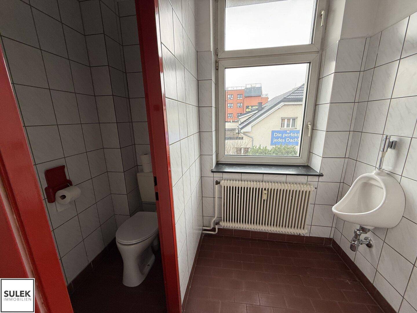 Badezimmer mit roten Fliesen, weißen Wandfliesen, Toilette, Urinal und Fenster.