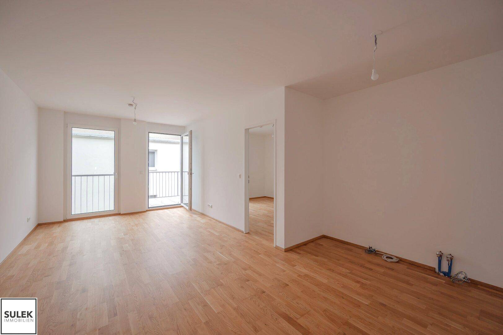 Geräumiges, leeres Wohnzimmer mit hellem Parkettboden und Zugang zu einem Balkon.