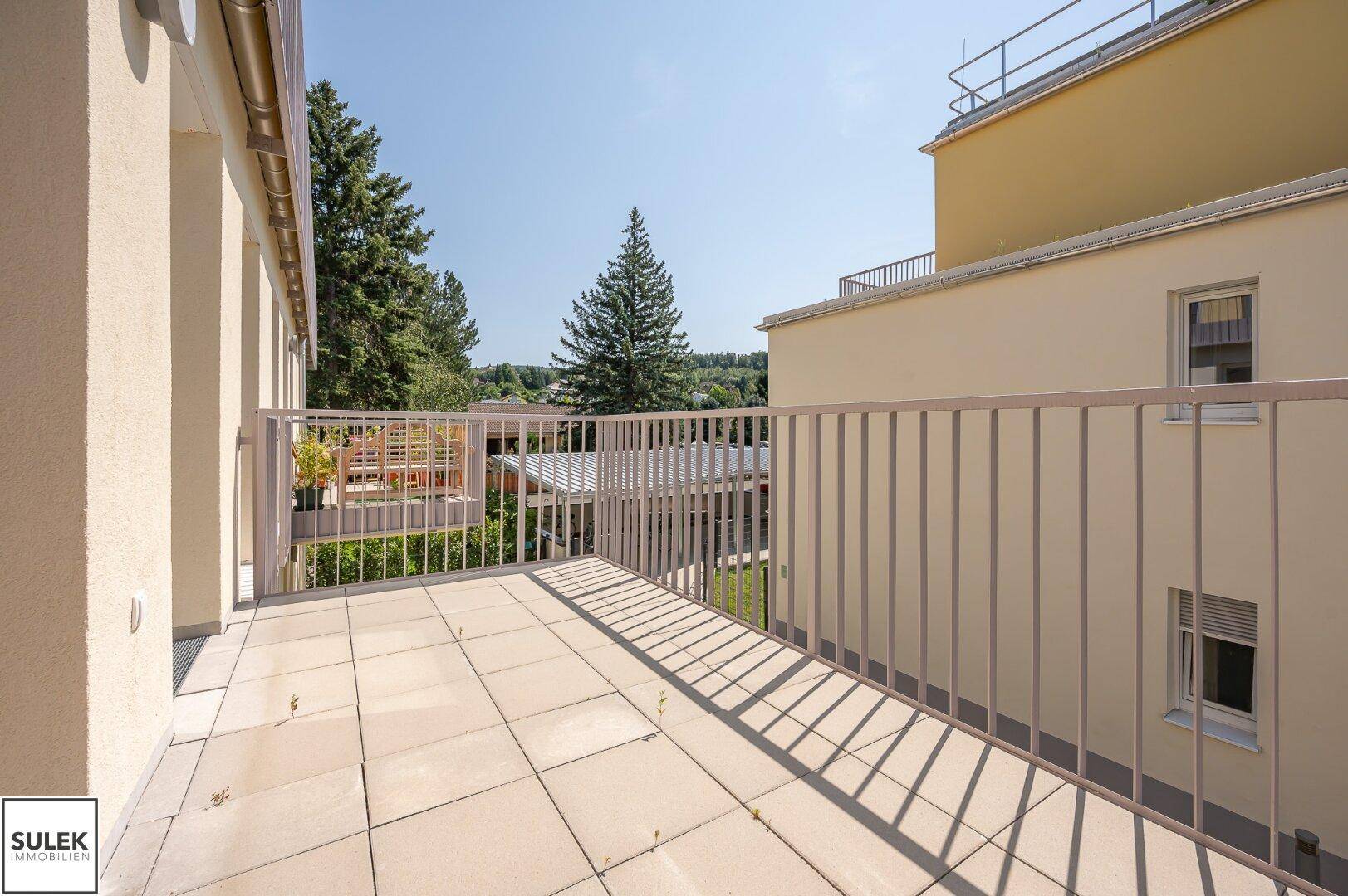 Sonniger Balkon mit Fliesenboden und weitem Blick auf Bäume und die Umgebung.