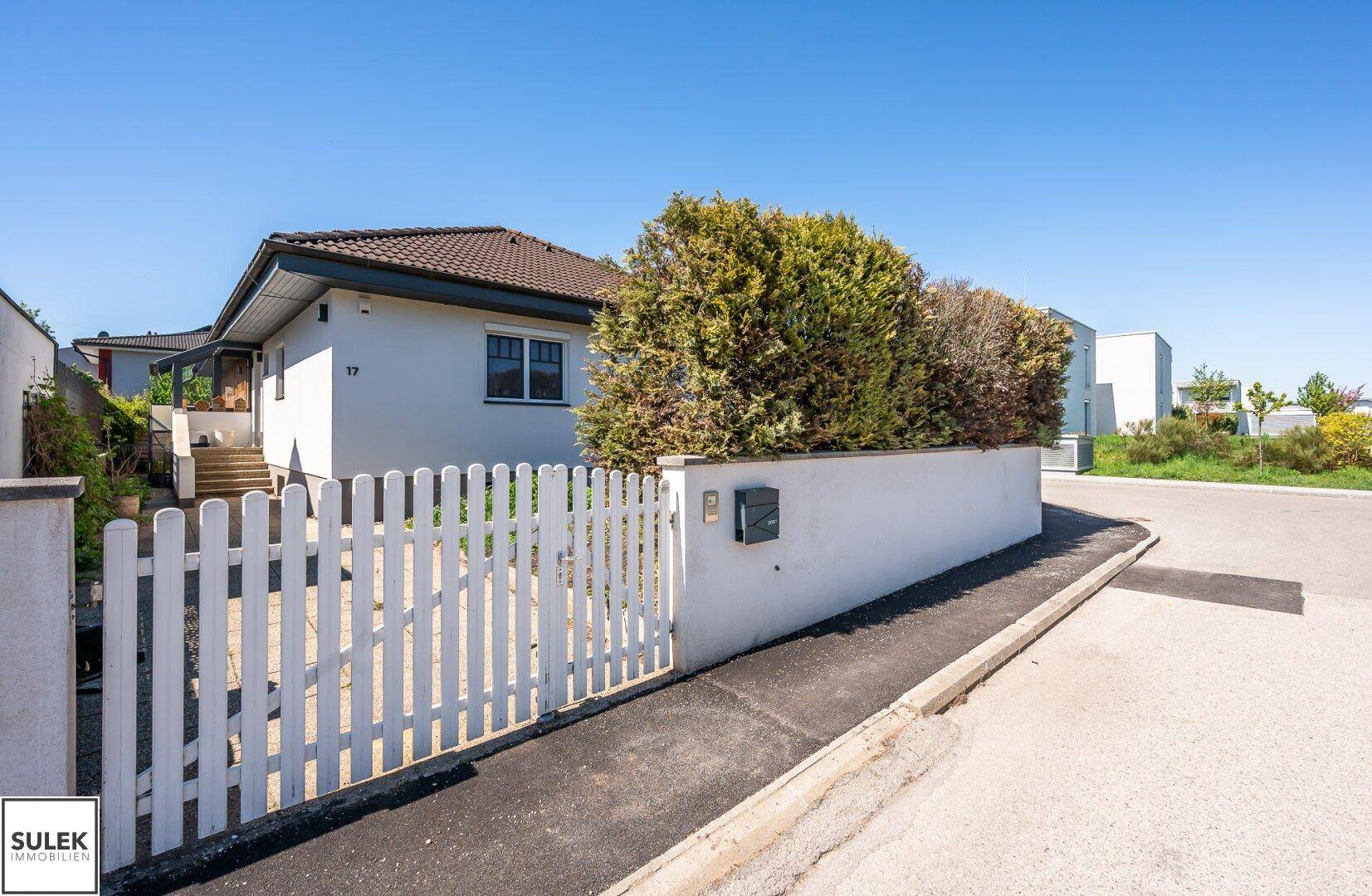 Einfamilienhaus mit weißem Lattenzaun und gepflegter Hecke an einer ruhigen Straße.