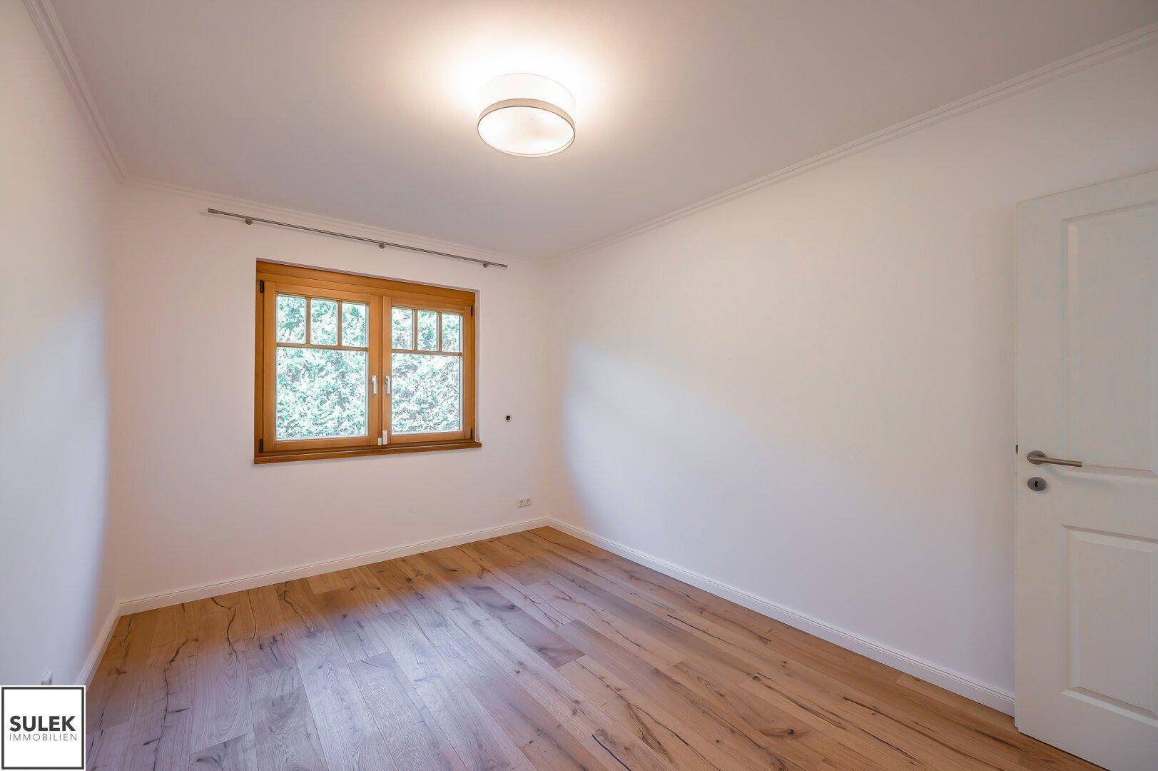 Freundliches Schlafzimmer mit Holzboden und einem Fenster, das einen schönen Ausblick ins Grüne ermöglicht.