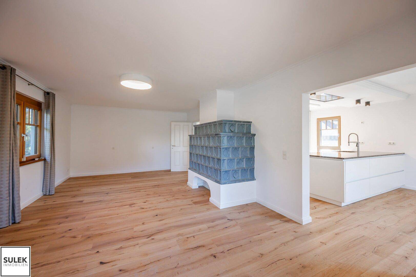 Offener Wohnbereich mit Kachelofen und angrenzender moderner Küche mit Insel, alles auf Holzboden.
