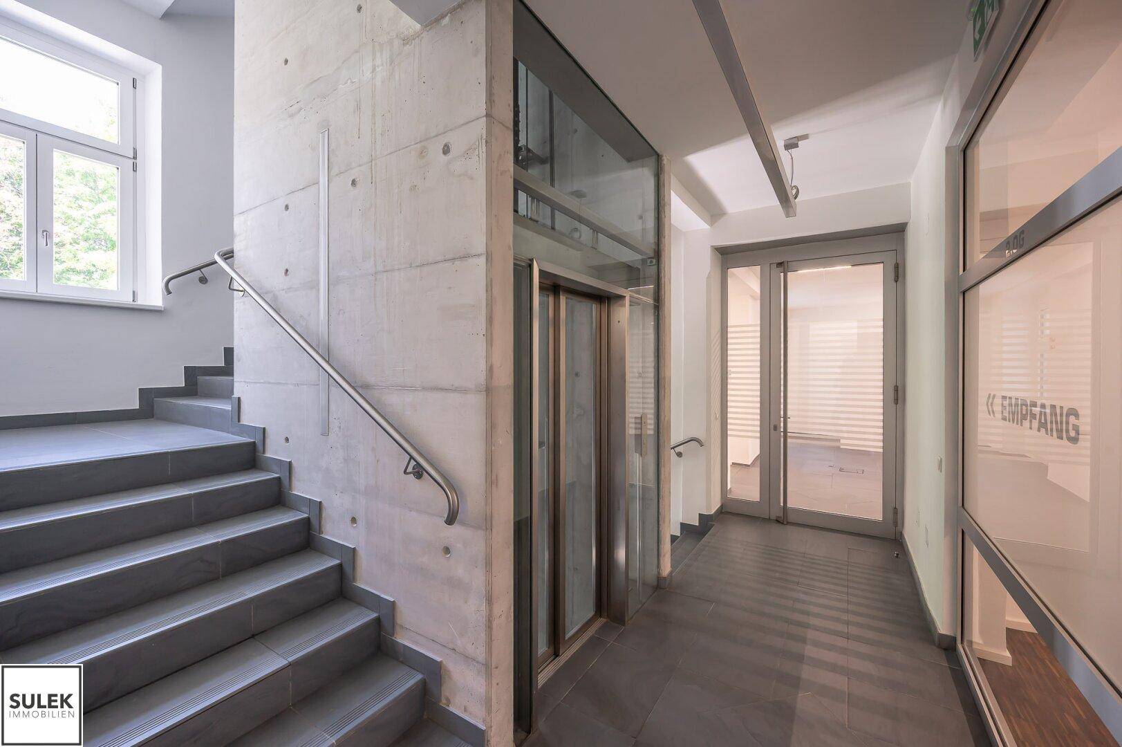Treppenhaus mit modernem Aufzug und Betonwänden, führt zu den Büroräumen und dem Empfangsbereich in einem zeitgemäßen Design.