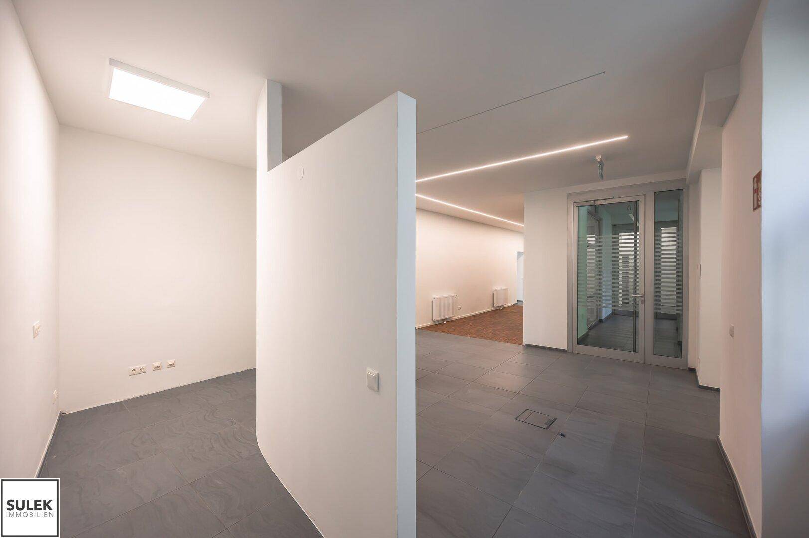 Offener Bürobereich mit geschwungener Wand, Fliesenboden und integrierter Beleuchtung, bietet flexible Gestaltungsmöglichkeiten für moderne Arbeitsplätze.