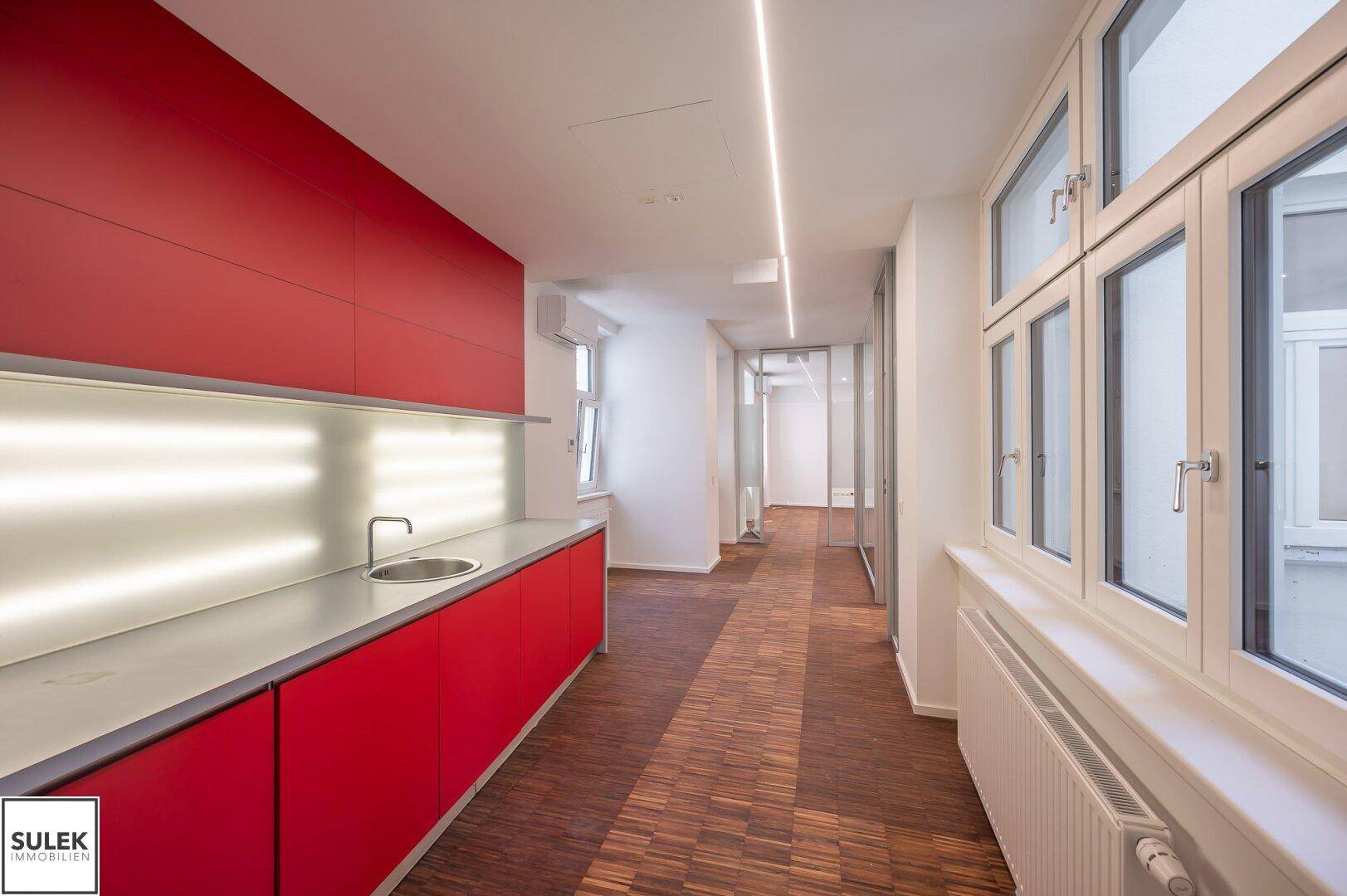 Moderne rote Küchenzeile mit Spüle und Unterschränken, integriert in einen hellen Bürobereich mit Holzboden und Fenstern.