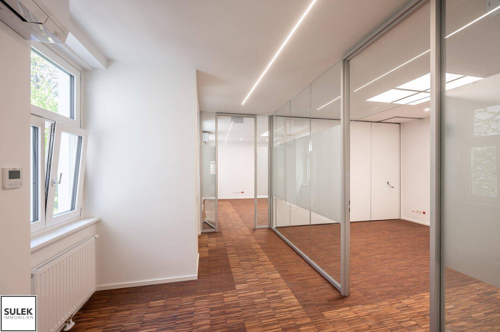 Heller Bürobereich mit Holzboden und Glaswänden, die für eine offene und lichtdurchflutete Atmosphäre sorgen.