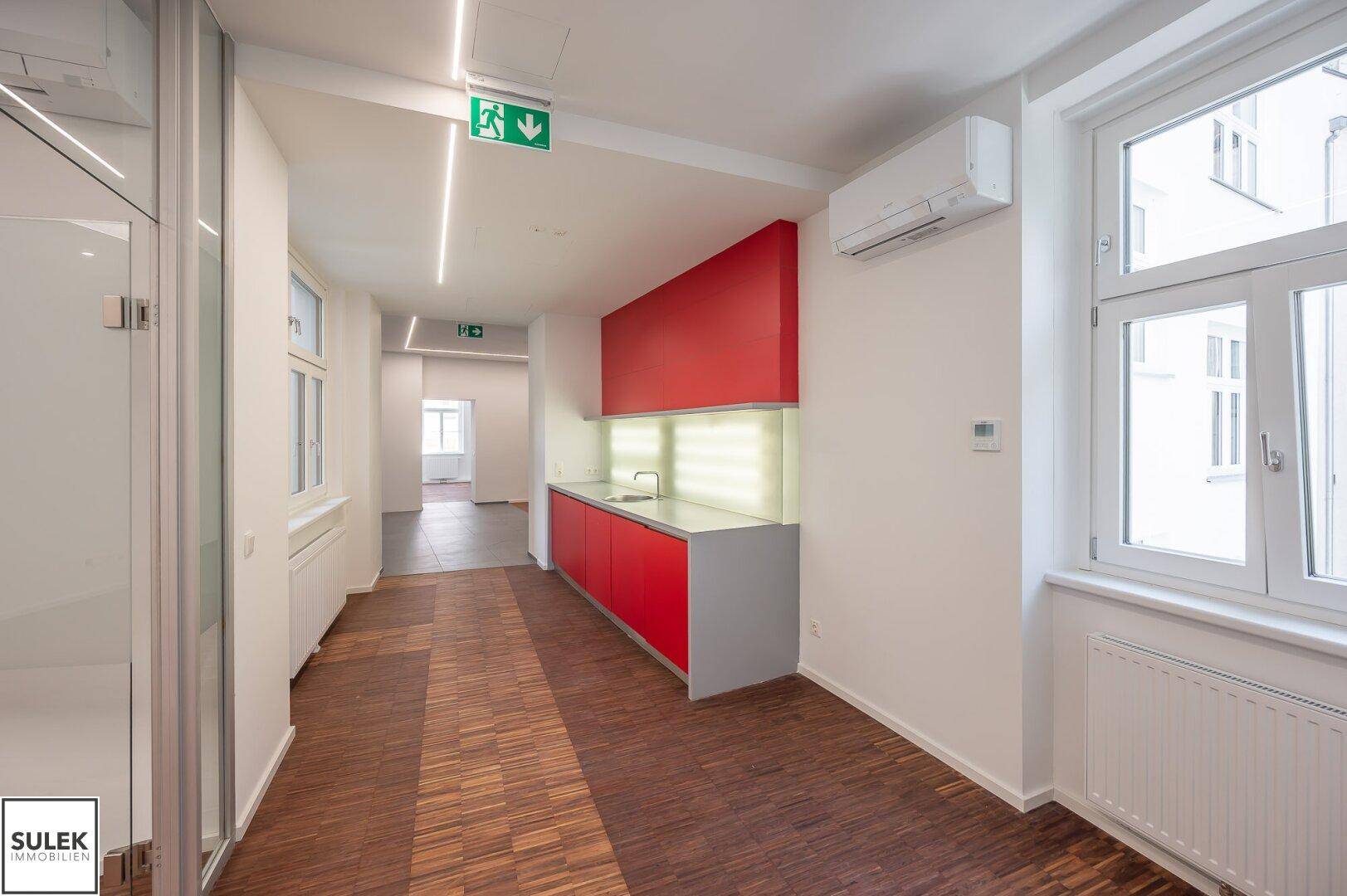 Funktionale rote Küchenzeile mit Spüle und viel Stauraum, in einem hellen Flur mit Holzboden und Fenstern.
