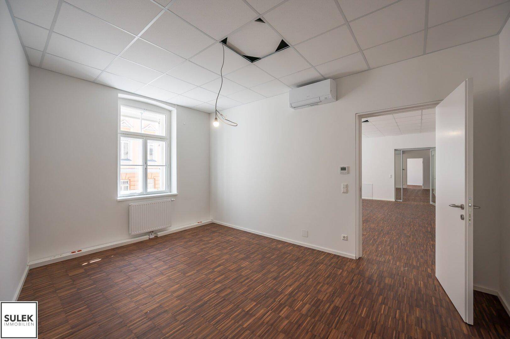 Helles Büro mit Holzboden und Rasterdecke, ausgestattet mit Klimaanlage und einem großen Fenster.