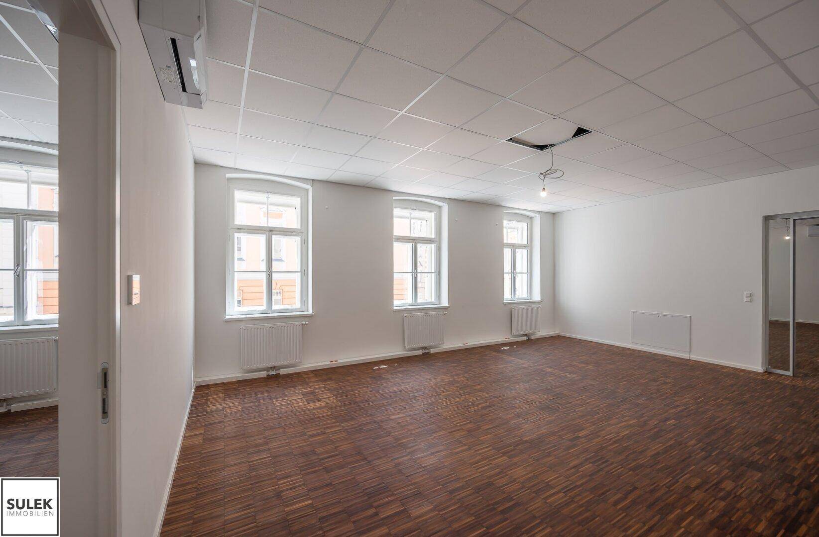Geräumiges Büro mit Holzboden und drei großen Fenstern, die für viel Tageslicht und eine angenehme Atmosphäre sorgen.