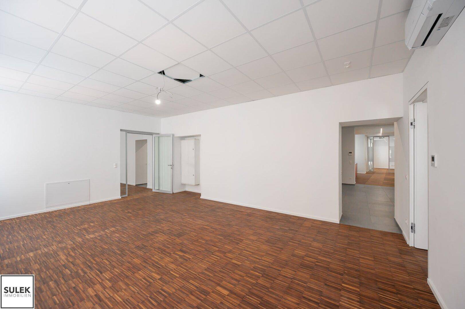 Großes Büro mit Holzboden und Rasterdecke, bietet viel Platz und ist durch mehrere Türen zugänglich.