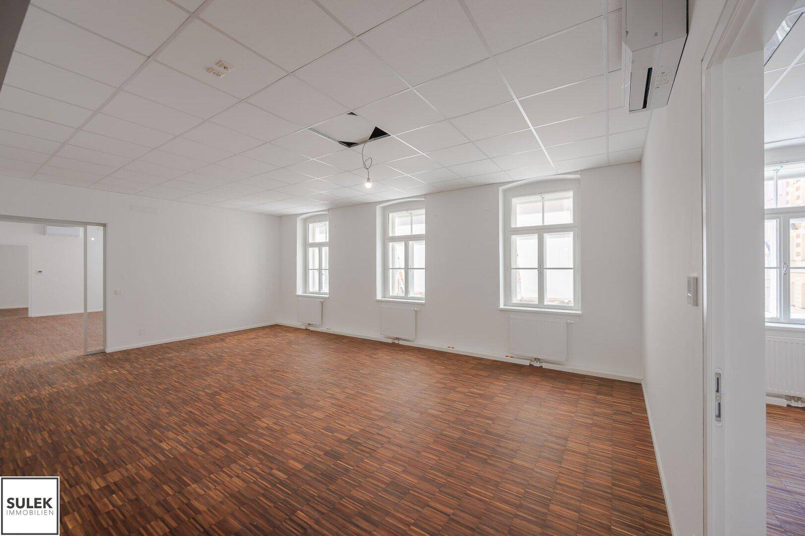 Helles Büro mit Holzboden und drei Fenstern, die eine gute Belichtung und Ausblick bieten.