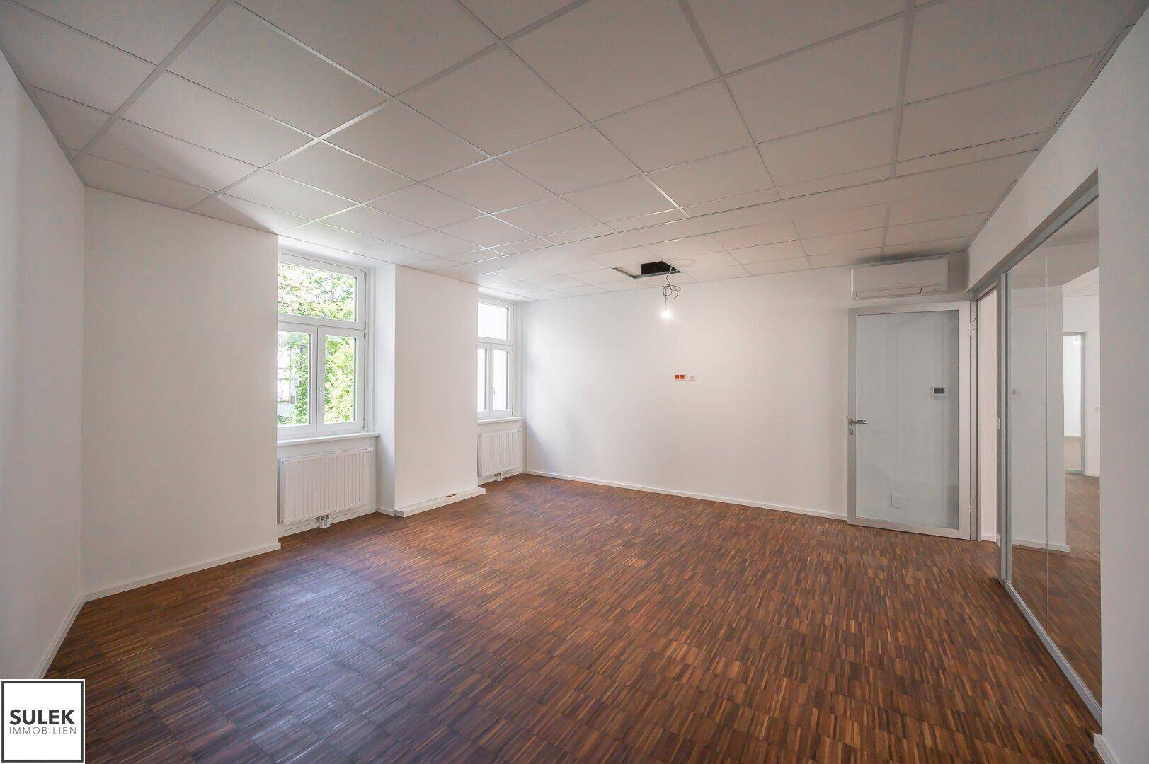 Helles Büro mit Holzboden und Rasterdecke, ausgestattet mit Klimaanlage und einem großen Fenster.
