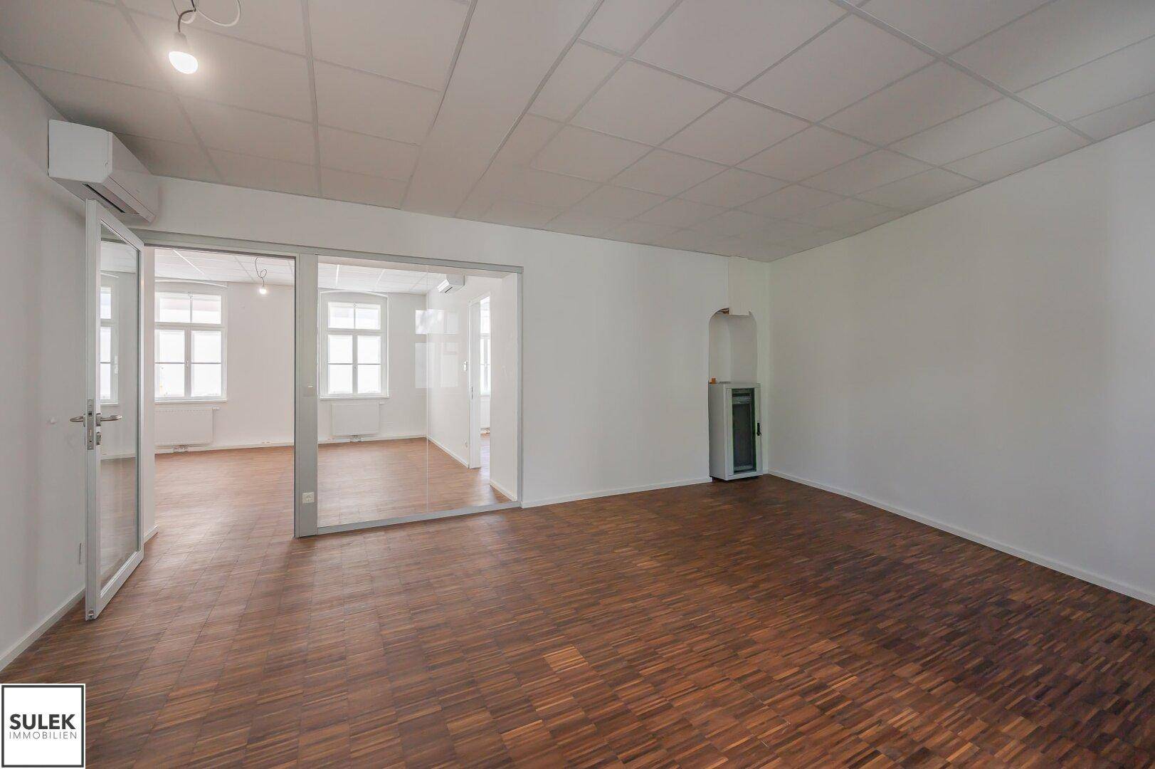 Geräumiges Büro mit Holzboden und Glaswänden, die eine offene und moderne Arbeitsumgebung schaffen, inklusive Kaminofen.