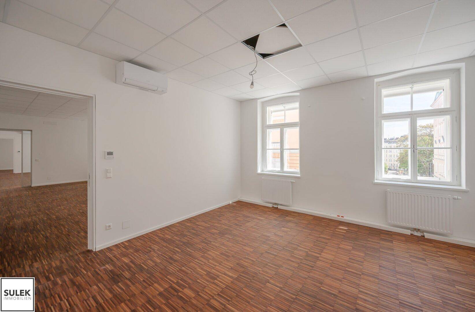 Helles Büro mit Holzboden und Rasterdecke, ausgestattet mit Klimaanlage und einem großen Fenster.