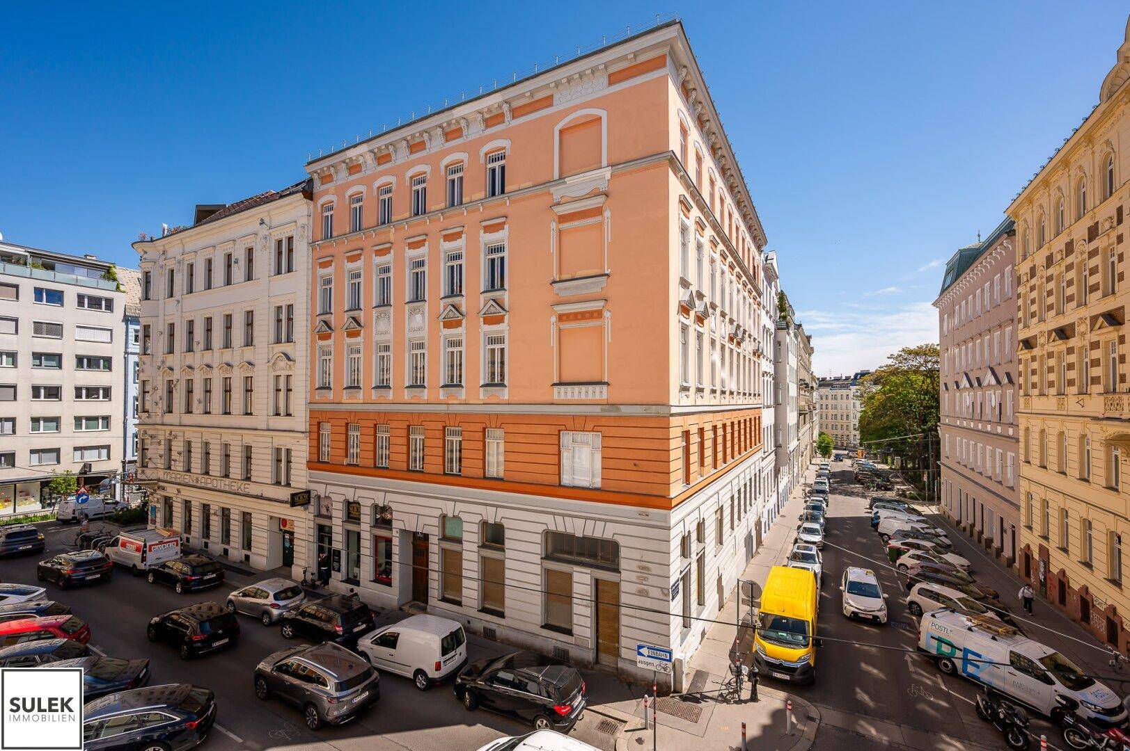 Historisches Gebäude mit oranger Fassade und vielen Fenstern, umgeben von einer belebten Stadtstraße unter blauem Himmel.