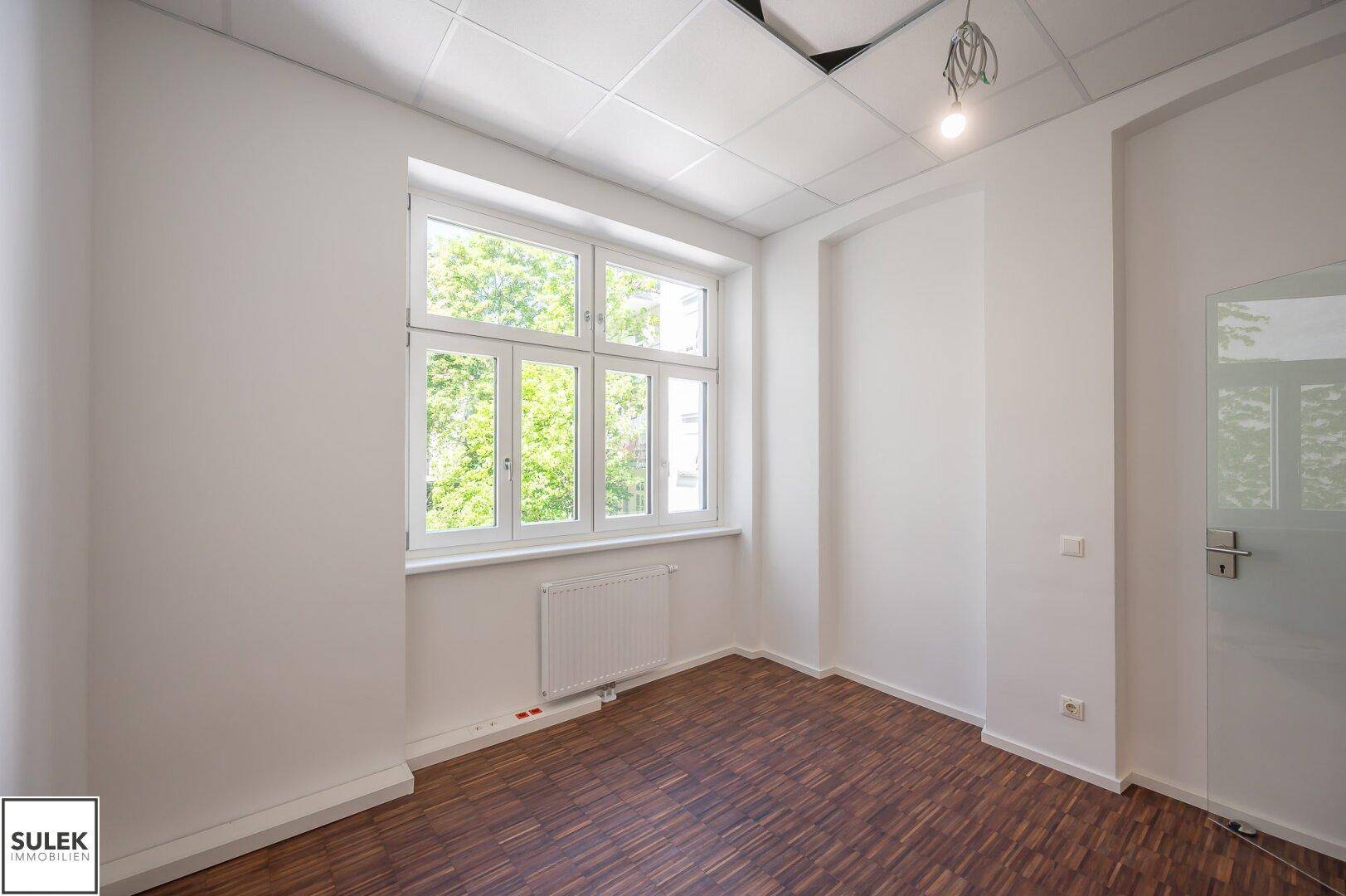 Helles Büro mit Holzboden und Rasterdecke, bietet einen Ausblick ins Grüne durch das große Fenster.