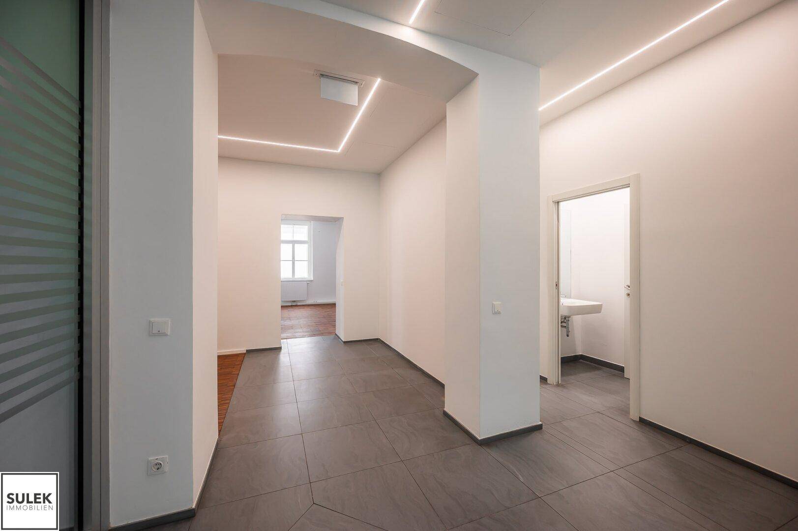 Heller Flur mit Fliesenboden und integrierter Beleuchtung, führt zu den Toiletten und weiteren Räumen.