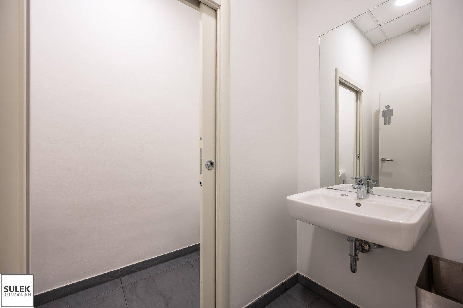 Heller Toilettenraum mit Waschbecken und großem Spiegel, bietet eine saubere und funktionale Ausstattung.