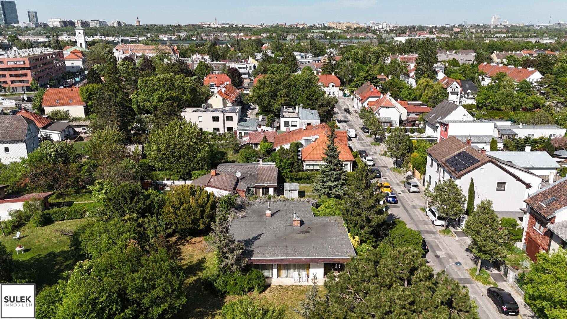 Panoramablick über die grüne Wohnsiedlung mit Einfamilienhäusern und städtischem Horizont unter blauem Himmel.