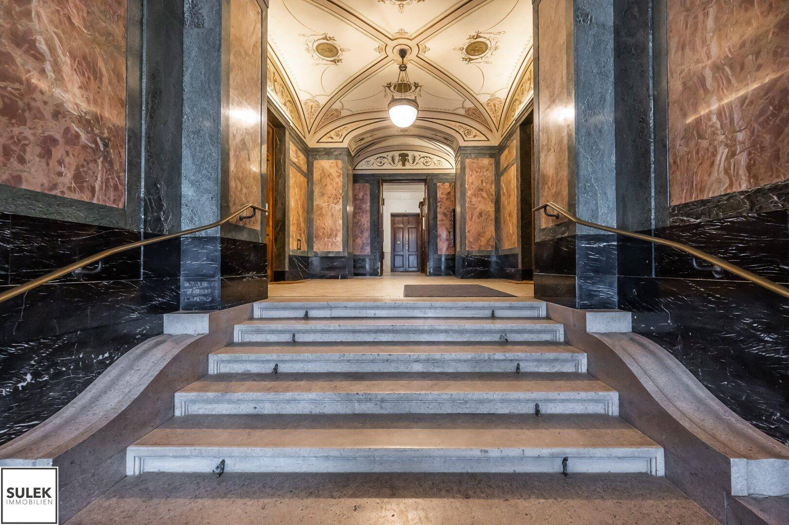 Repräsentativer Eingangsbereich mit breiter Treppe, Marmorwänden und dekorativer Decke.