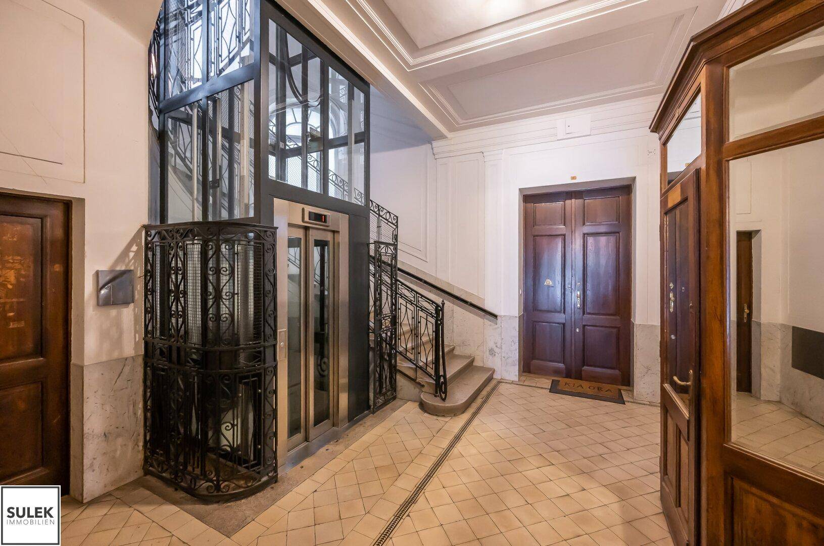Elegantes Treppenhaus mit historischem Aufzug und verzierten Decken in einem Altbau.